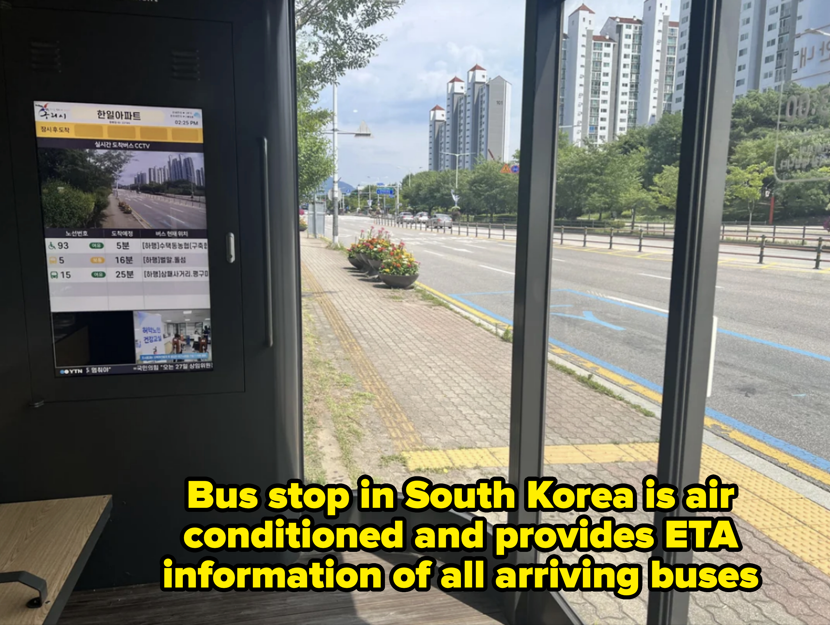 Bus stop interior with digital screen showing bus schedule and street view with tall buildings and trees in the background