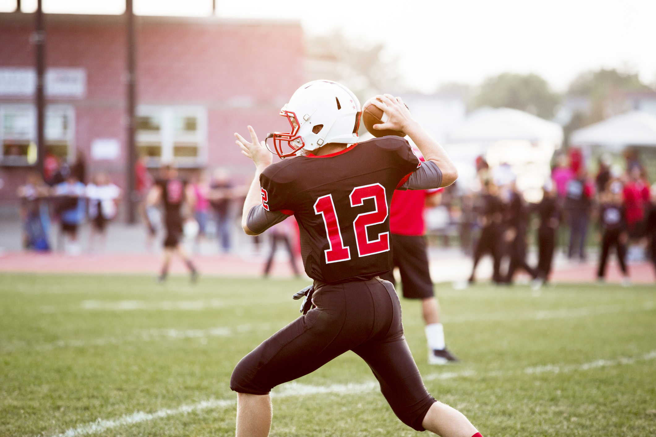 A high school football quarterback wearing a uniform and helmet, number 12, is poised to throw the ball on a grassy football field during a game