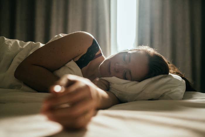 A woman is peacefully sleeping on a bed, lying on her side with a hand stretched out. Morning light softly illuminates the room
