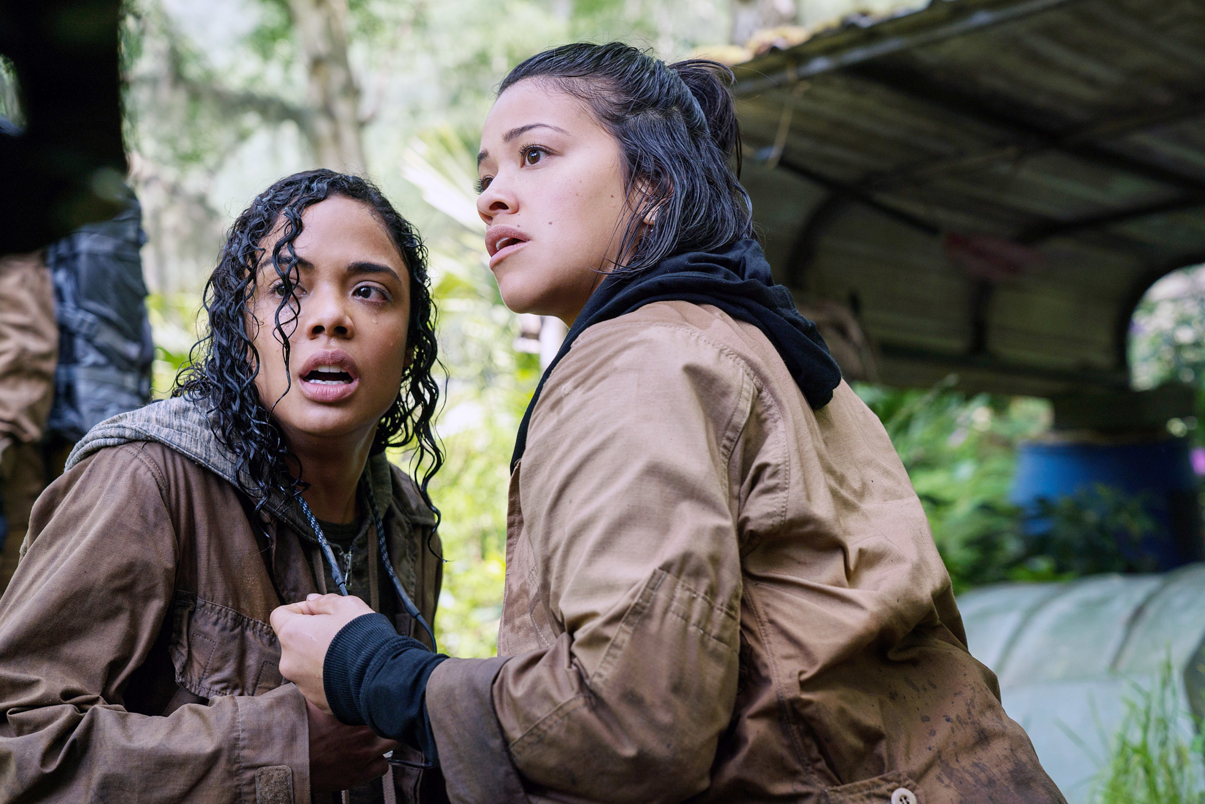 Gina Rodriguez and Tessa Thompson, both wearing brown jackets, appear startled in a jungle-like setting in this gripping scene