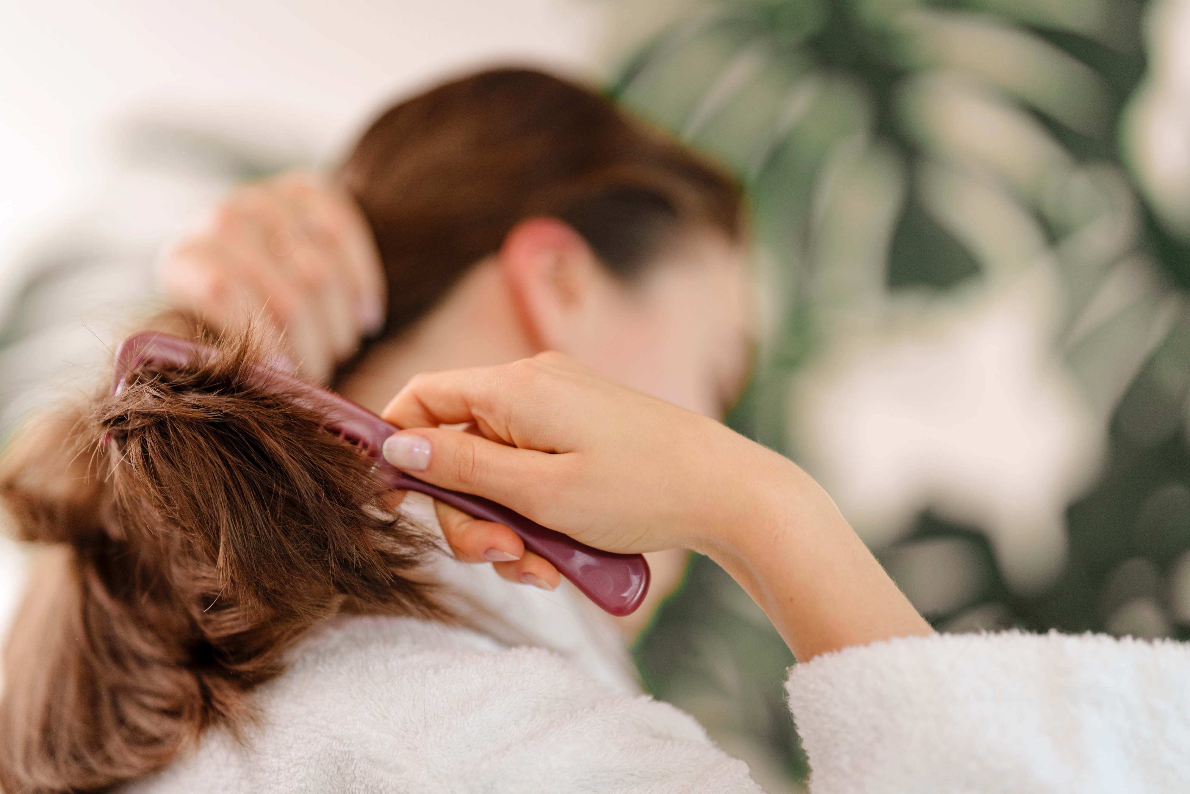 A person in a white robe brushing their brown hair, showing a close-up of their hands and head. Large green plant is in the blurred background