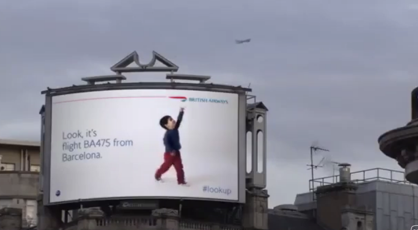 British Airways billboard shows a child pointing to a plane in the sky with text: "Look, it's flight BA475 from Barcelona." #lookup