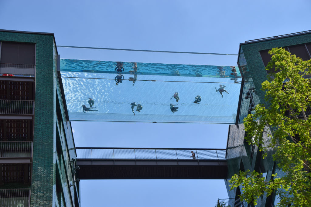 People swim and lounge in a glass-bottom pool suspended between two buildings high above the ground, with a person walking on a bridge below