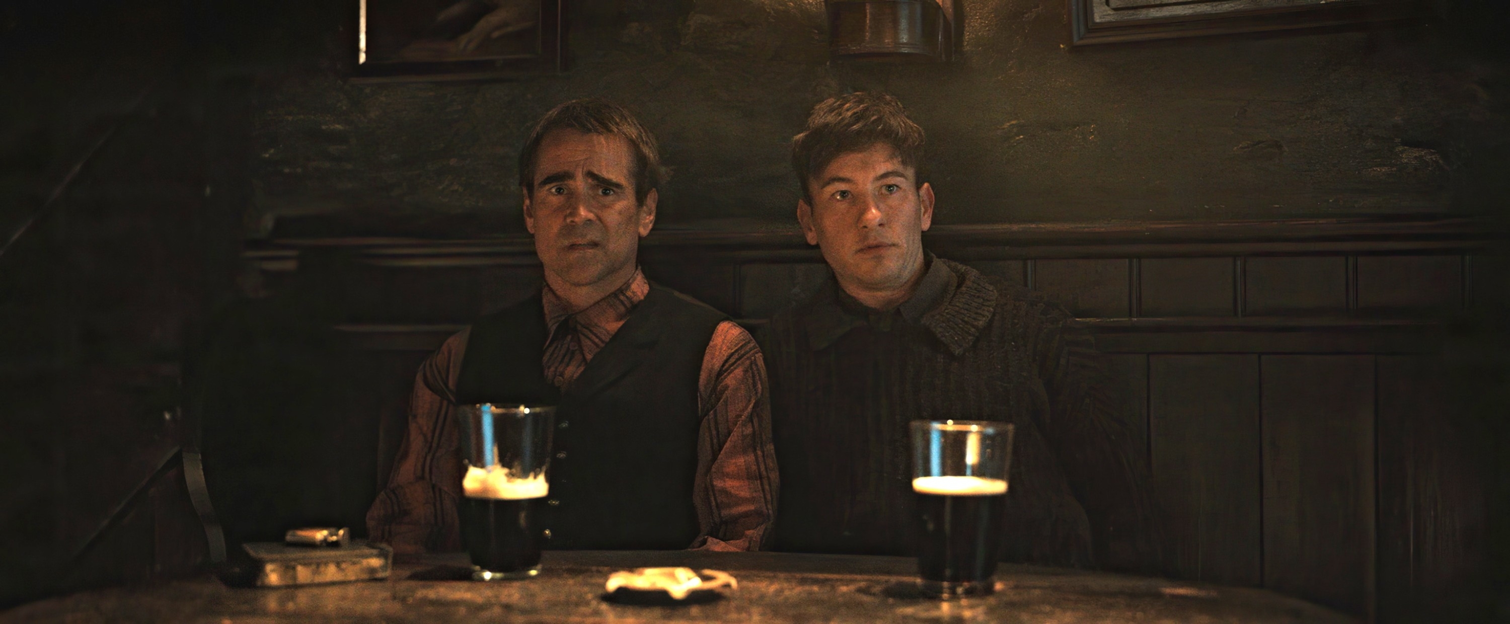 Colin Farrell and Barry Keoghan sit at a wooden table in a dimly lit pub, each with a half-full pint of beer in front of them
