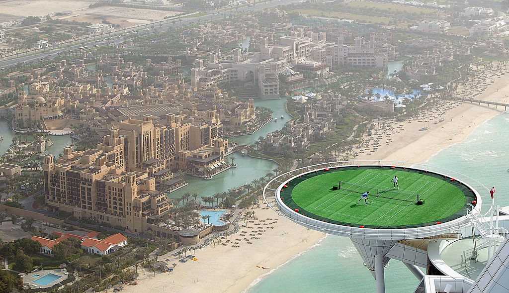 Aerial view of a helipad turned tennis court on a tall building, with people playing tennis. The backdrop is a coastal resort with an expansive beach and luxurious buildings
