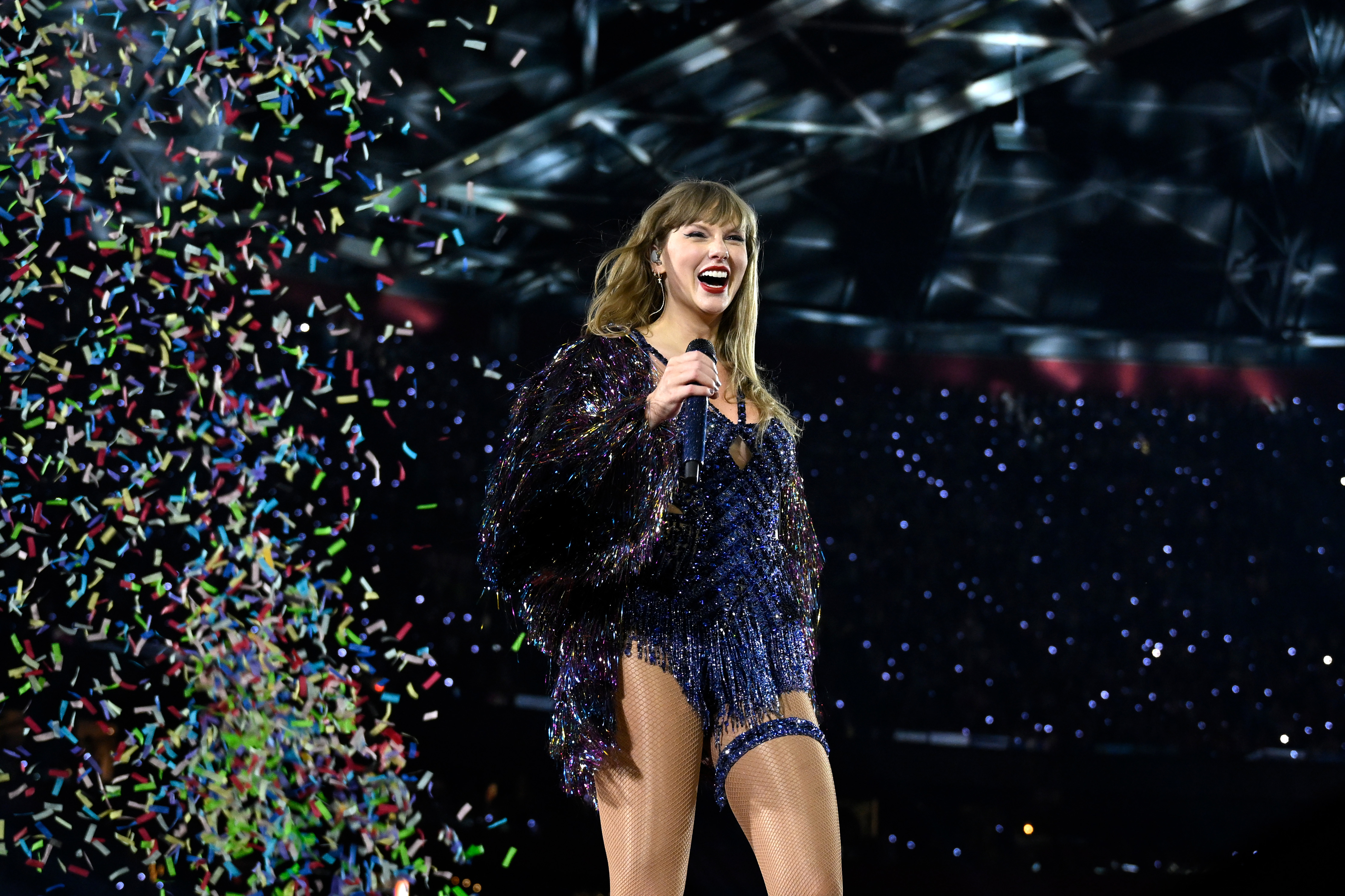 Taylor Swift on stage, smiling and holding a microphone, wearing a sparkling outfit with fringe. Confetti is falling around her during a live performance
