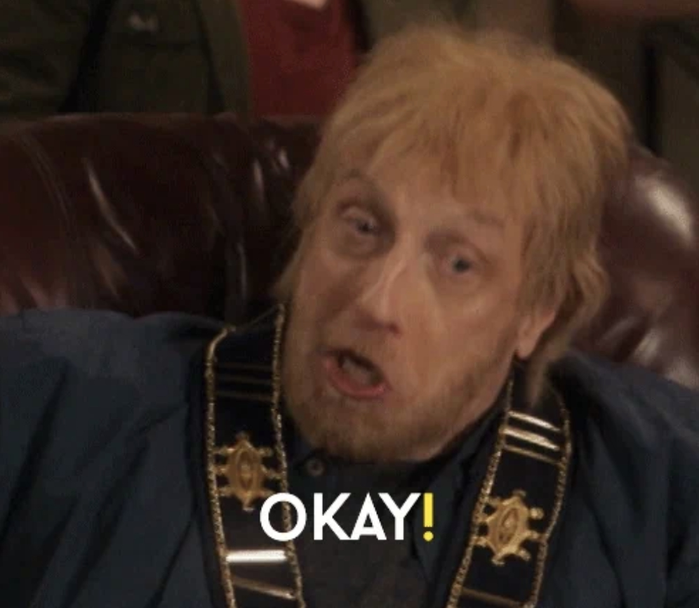 Man in a blue jacket with a gold chain, sitting and yelling "OKAY!"