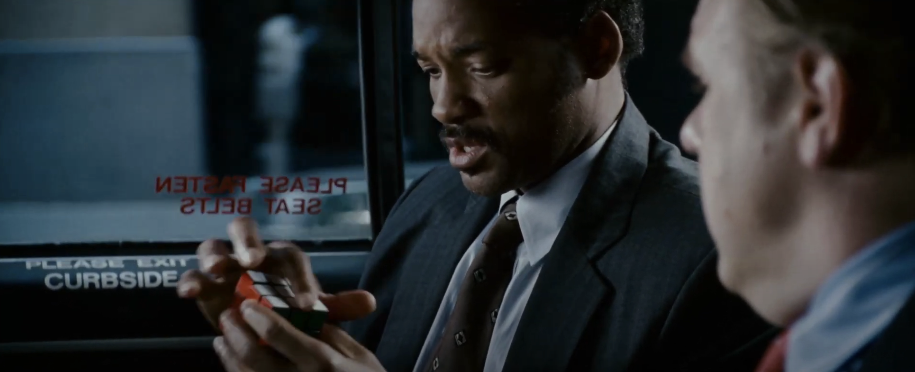 Will Smith and another actor are sitting in a car, with Will Smith holding and solving a Rubik's Cube intently