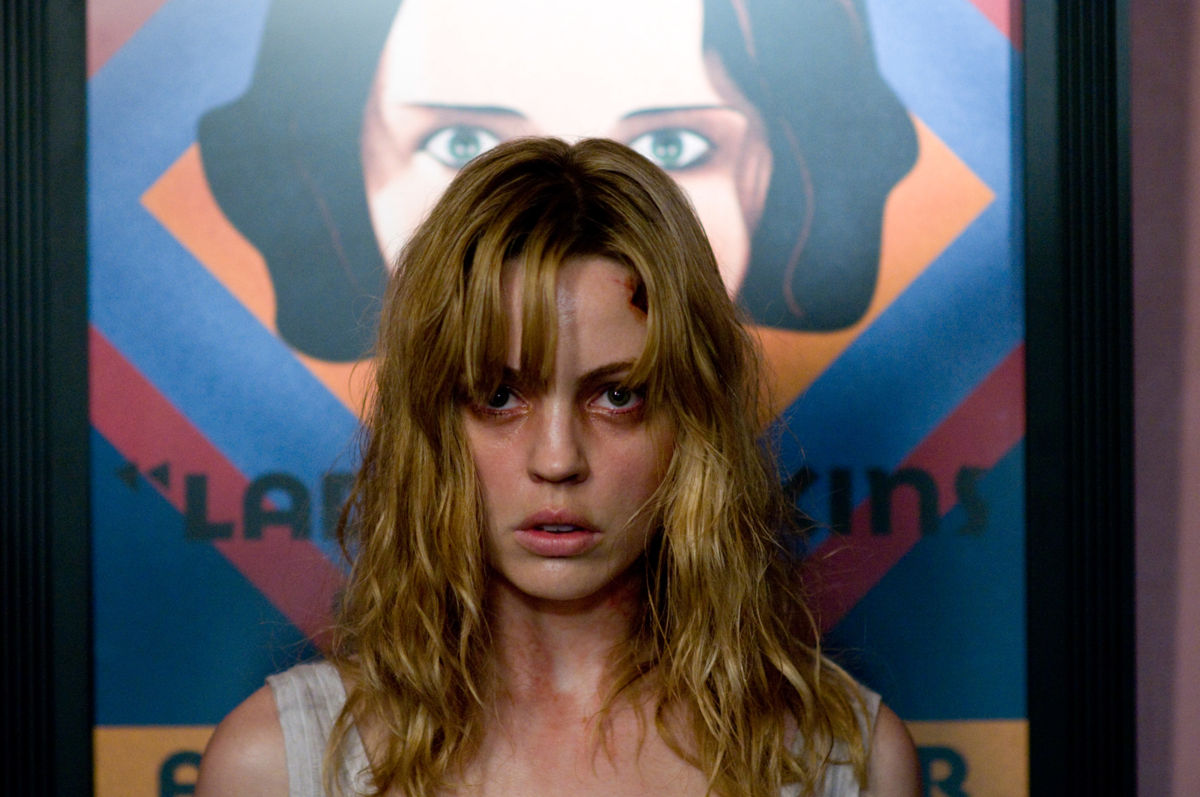 Amy Smart is standing in front of a poster featuring a woman's face, with a bruised look and disheveled hair