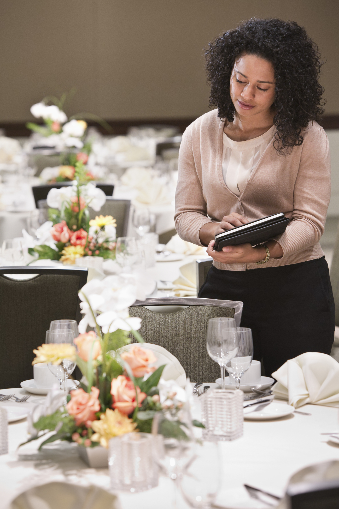 A woman reviews notes on a tablet while standing beside elegantly set tables with floral centerpieces, prepared for a wedding reception