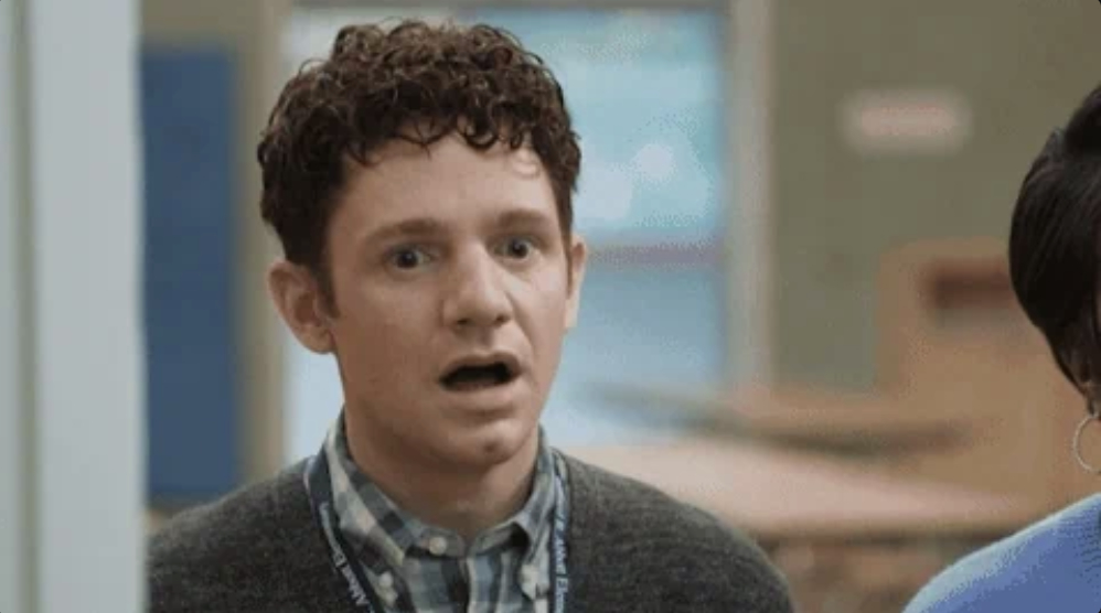 A young man with curly hair and a surprised expression stands indoors, wearing a checkered shirt and a sweater