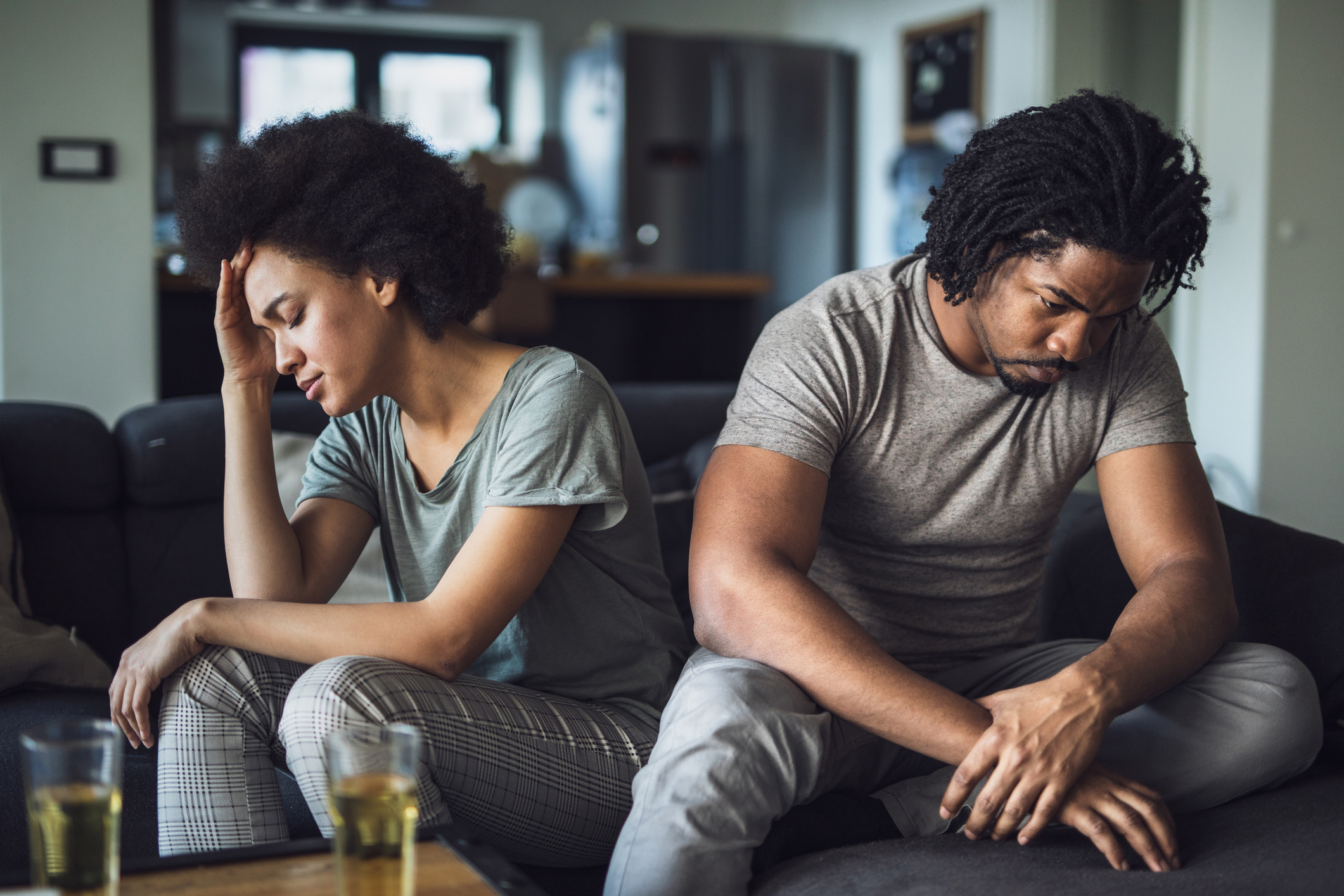 Two people sit on a couch, looking upset and avoiding eye contact. The woman has her hand on her forehead, while the man looks down with his hands on his knees
