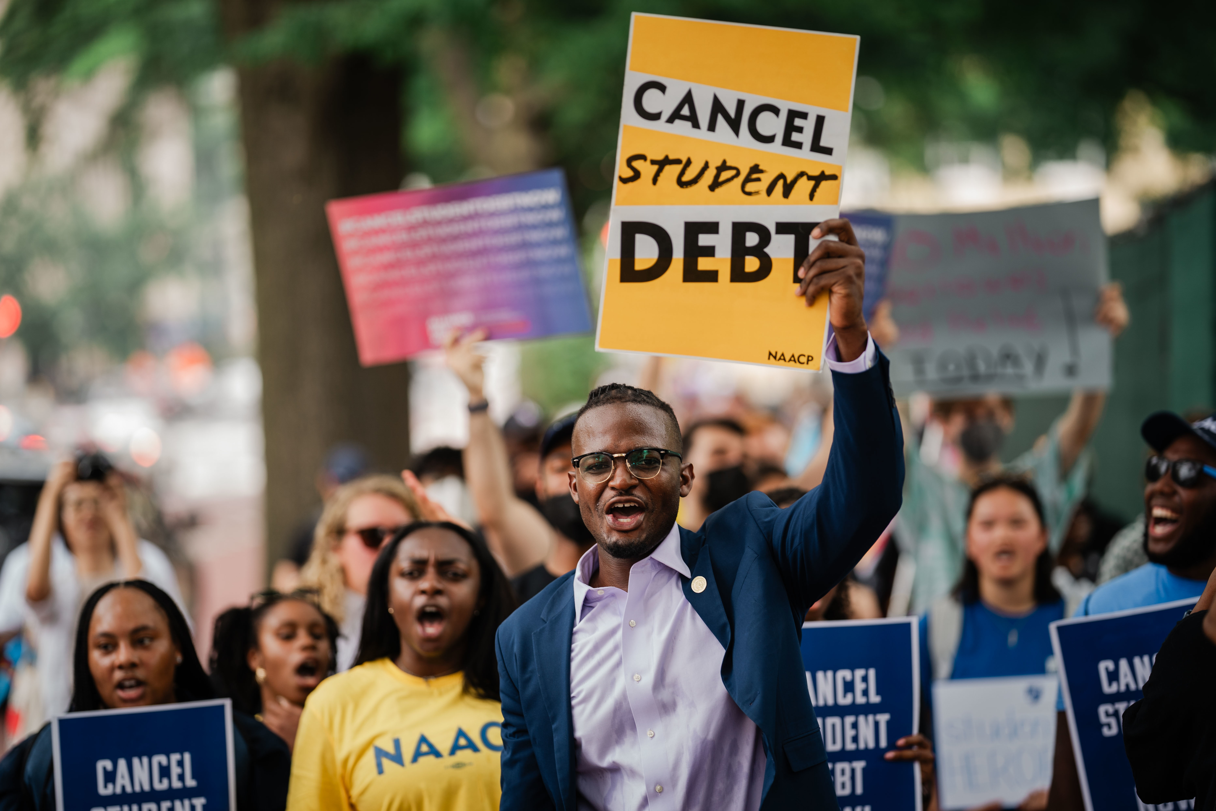 A group of protestors, led by a man holding a &quot;Cancel Student Debt&quot; sign, rally for student debt cancellation. Other participants hold signs and wear NAACP shirts