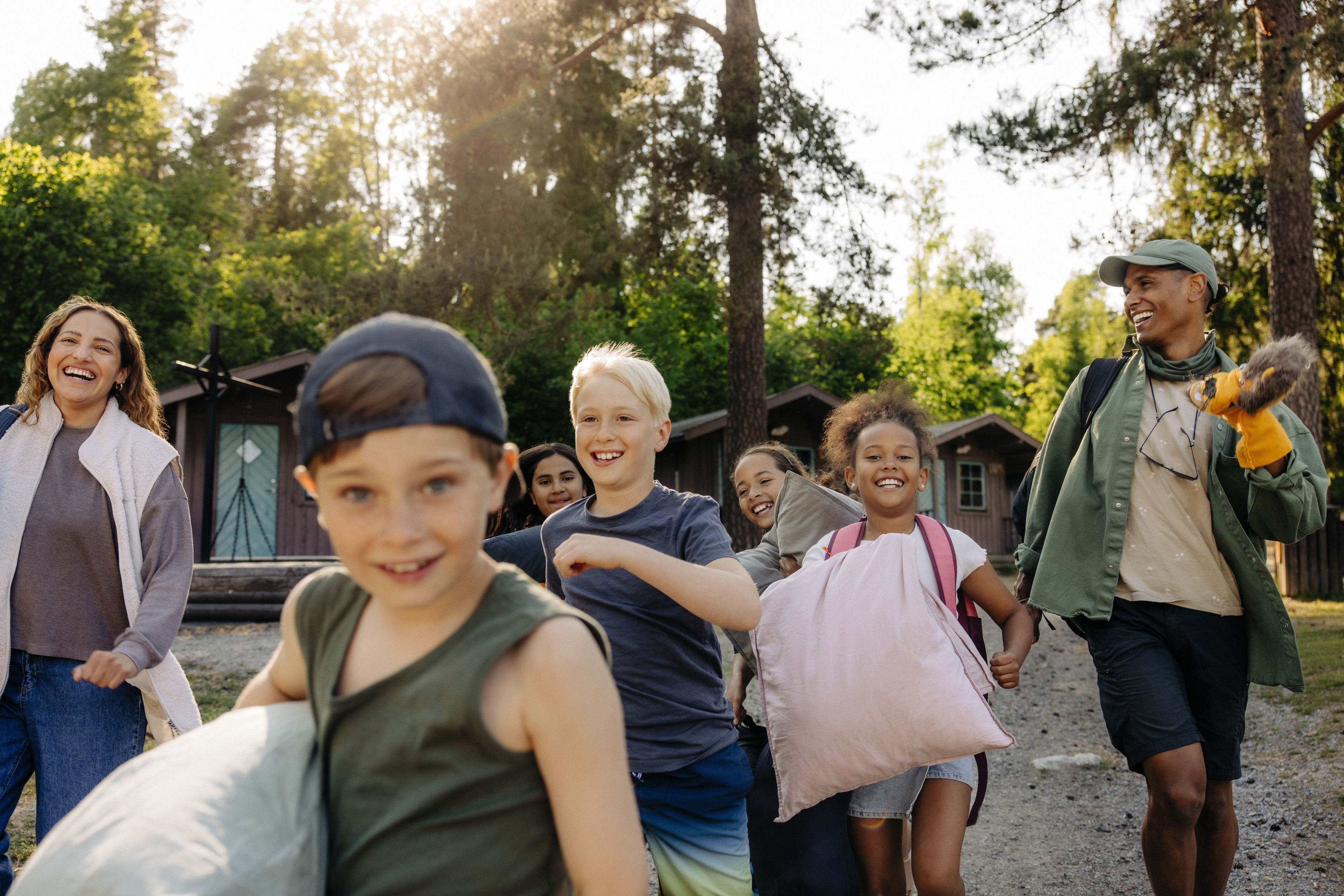 A group of children and an adult guide are smiling and running toward the camera, each holding pillows and backpacks, in an outdoor camp setting with cabins and trees