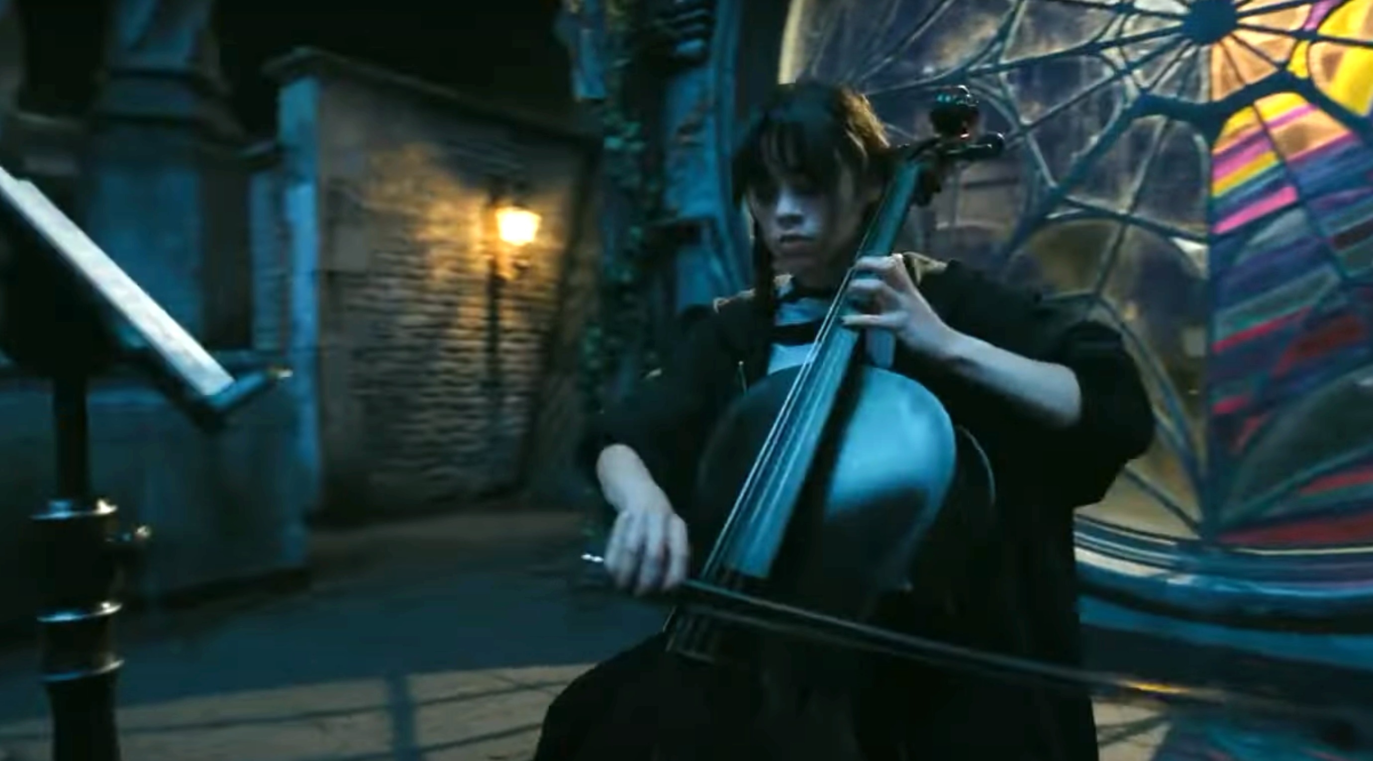 Jenna Ortega, dressed in a gothic-style outfit, plays the cello in front of a large, intricate stained-glass window in a scene from &quot;Wednesday.&quot;