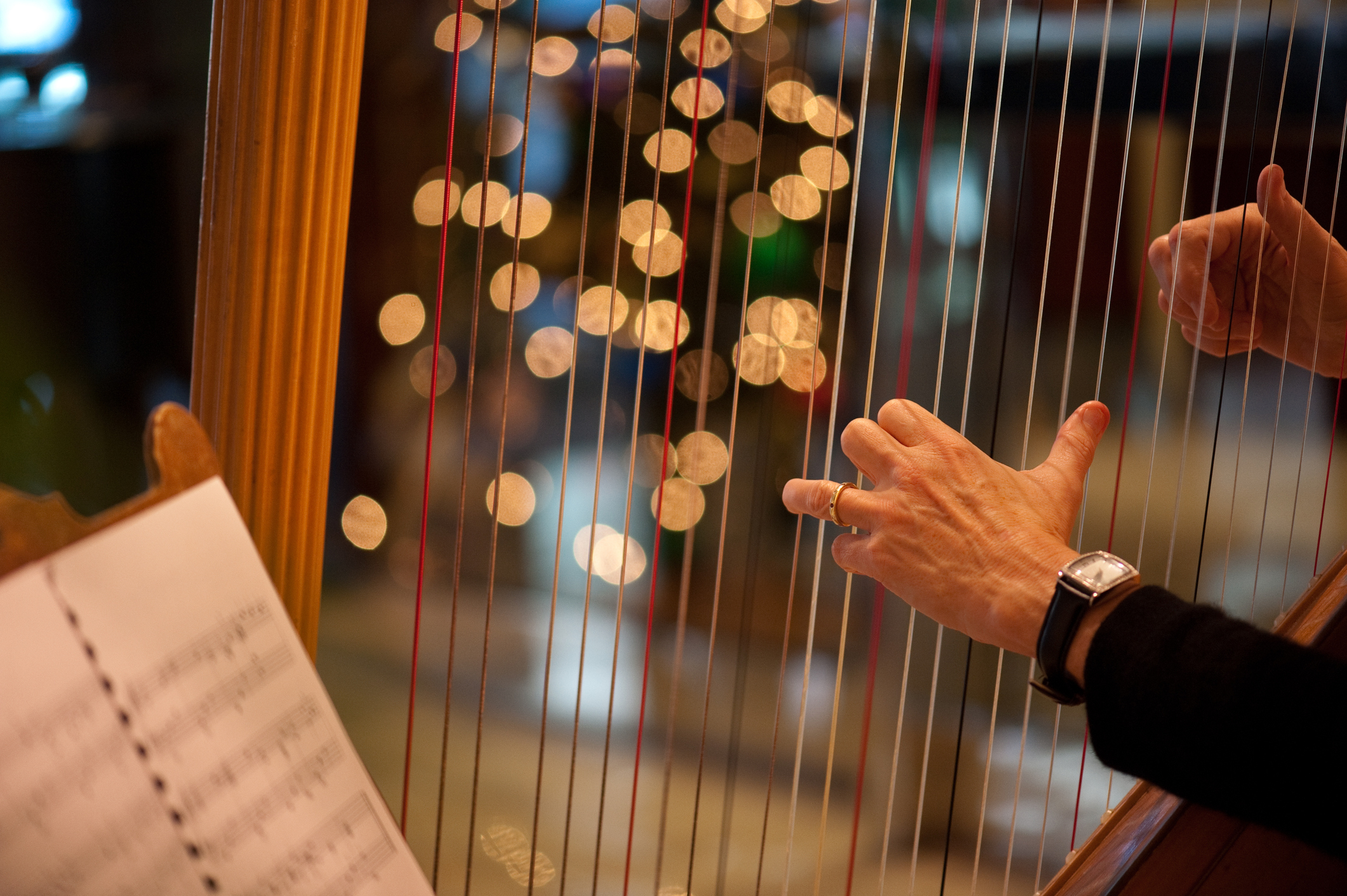 Close-up of a harpist's hands playing the instrument, with sheet music in the foreground and blurred lights in the background, possibly at a wedding