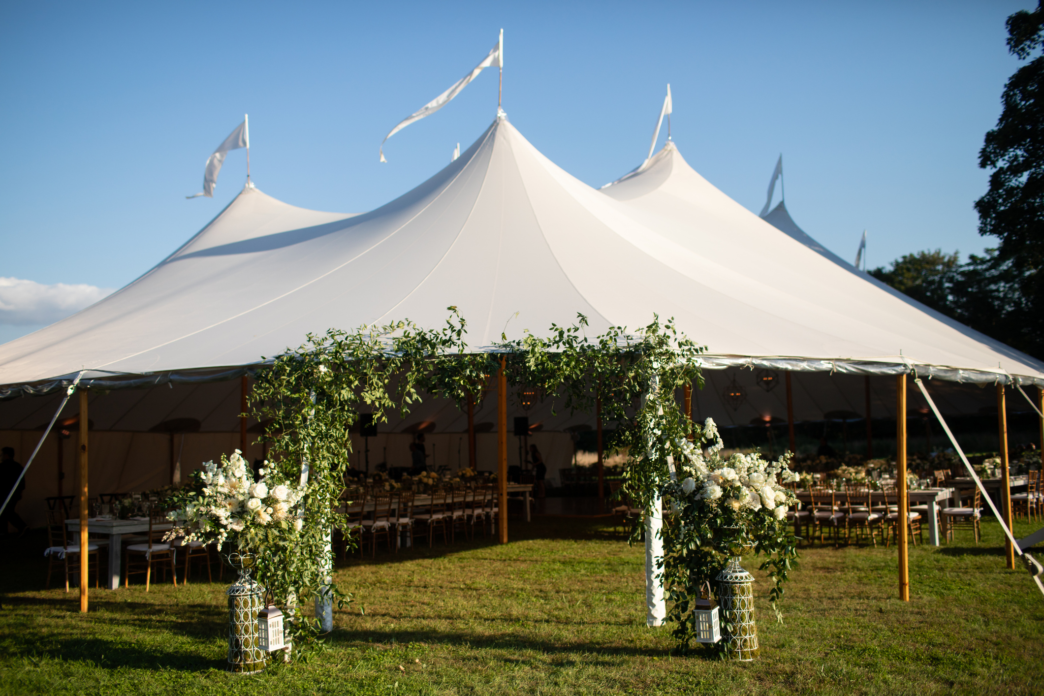 Outdoor wedding tent with flags on top, decorated archway with white flowers and greenery at the entrance, chairs and tables inside