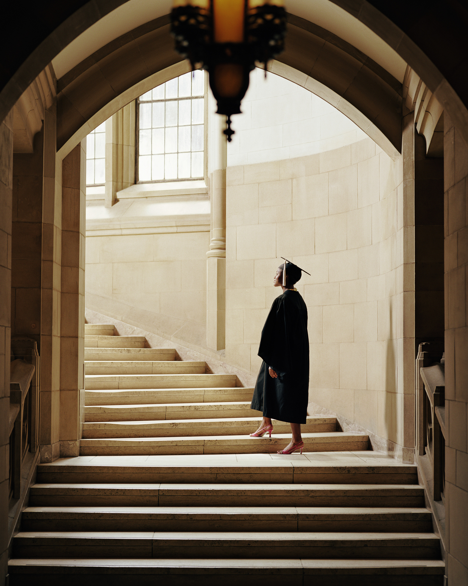 Person in graduation attire standing at the bottom of a grand staircase, looking up towards the light coming through large windows nearby
