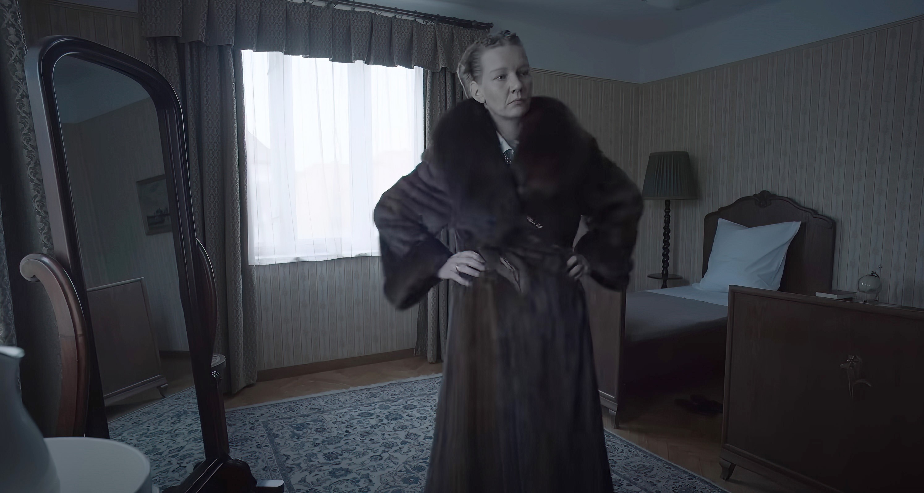 A person stands confidently in a vintage-styled bedroom, wearing a luxurious fur coat. A bed, lamp, and a large mirror are visible in the room