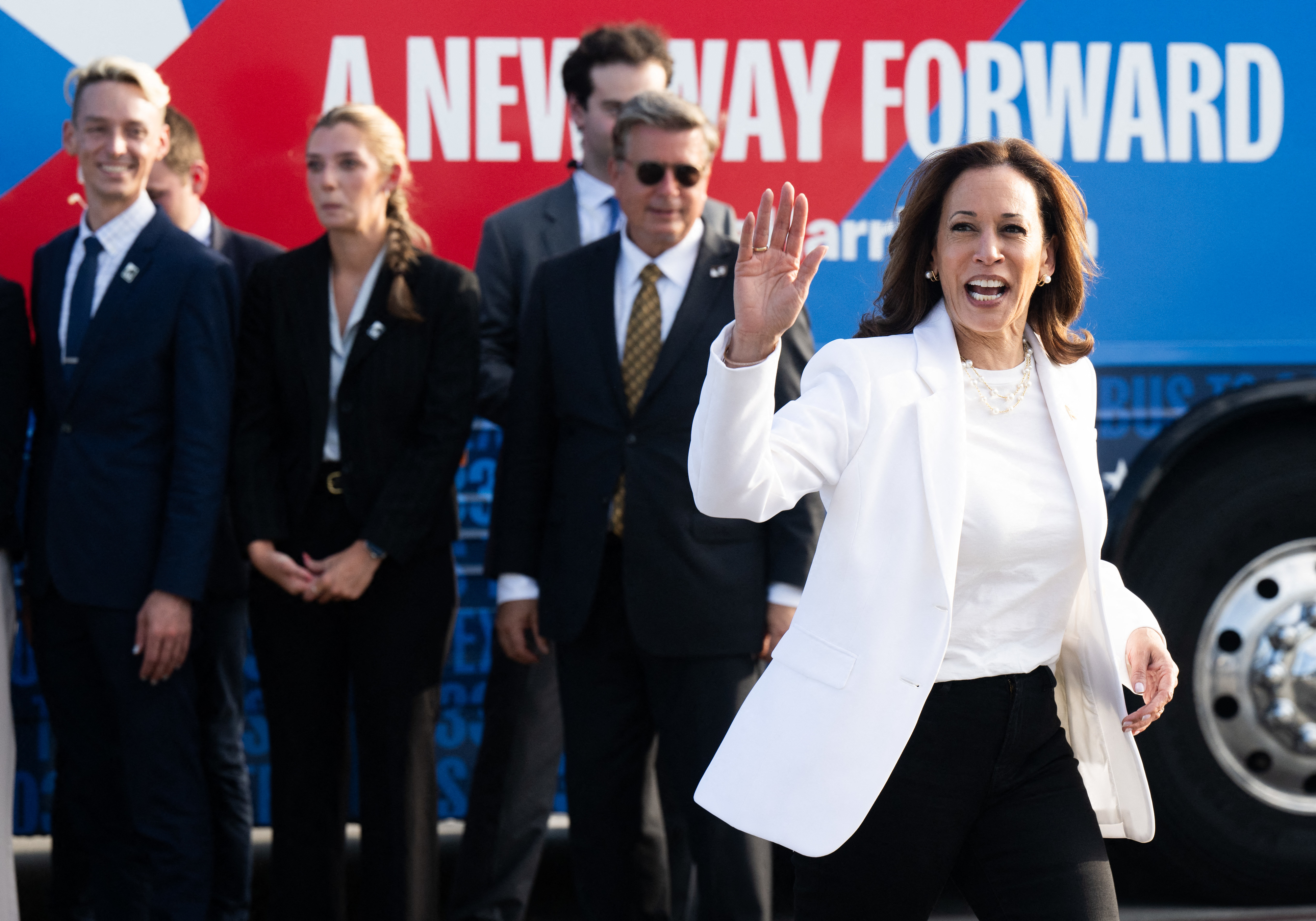 Kamala Harris waves while walking with a group of unidentified colleagues in front of a campaign bus with the text &quot;A New Way Forward.&quot;
