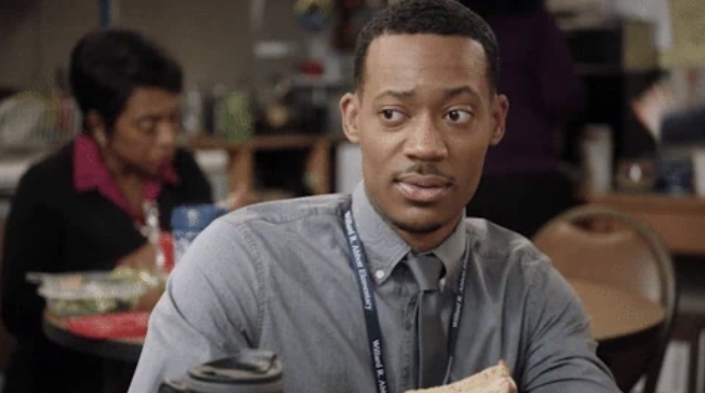 A man with a work badge around his neck holds a sandwich and looks to the side in a cafeteria setting