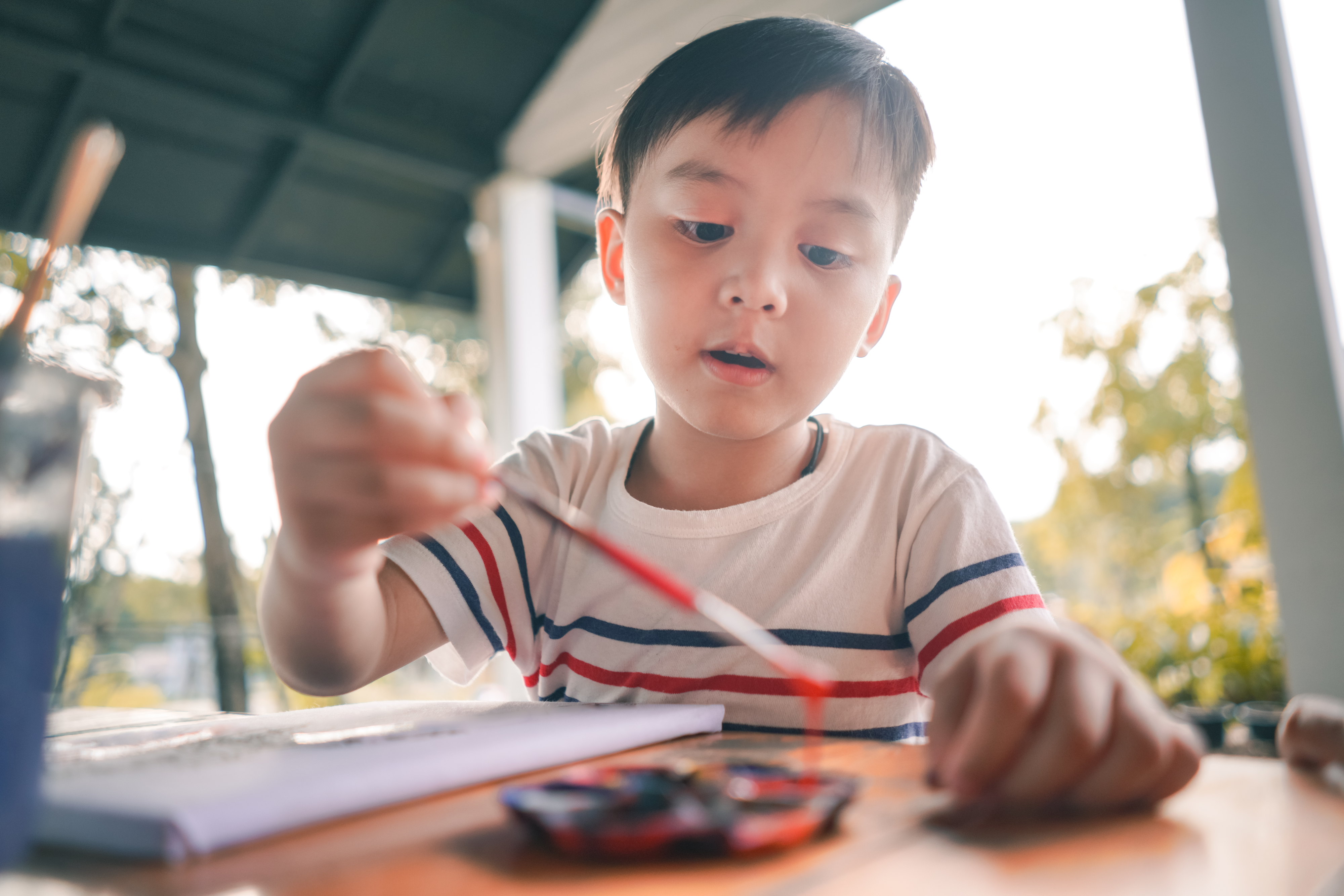 A young child wearing a striped t-shirt is painting with a brush at an outdoor table. Various painting supplies are on the table