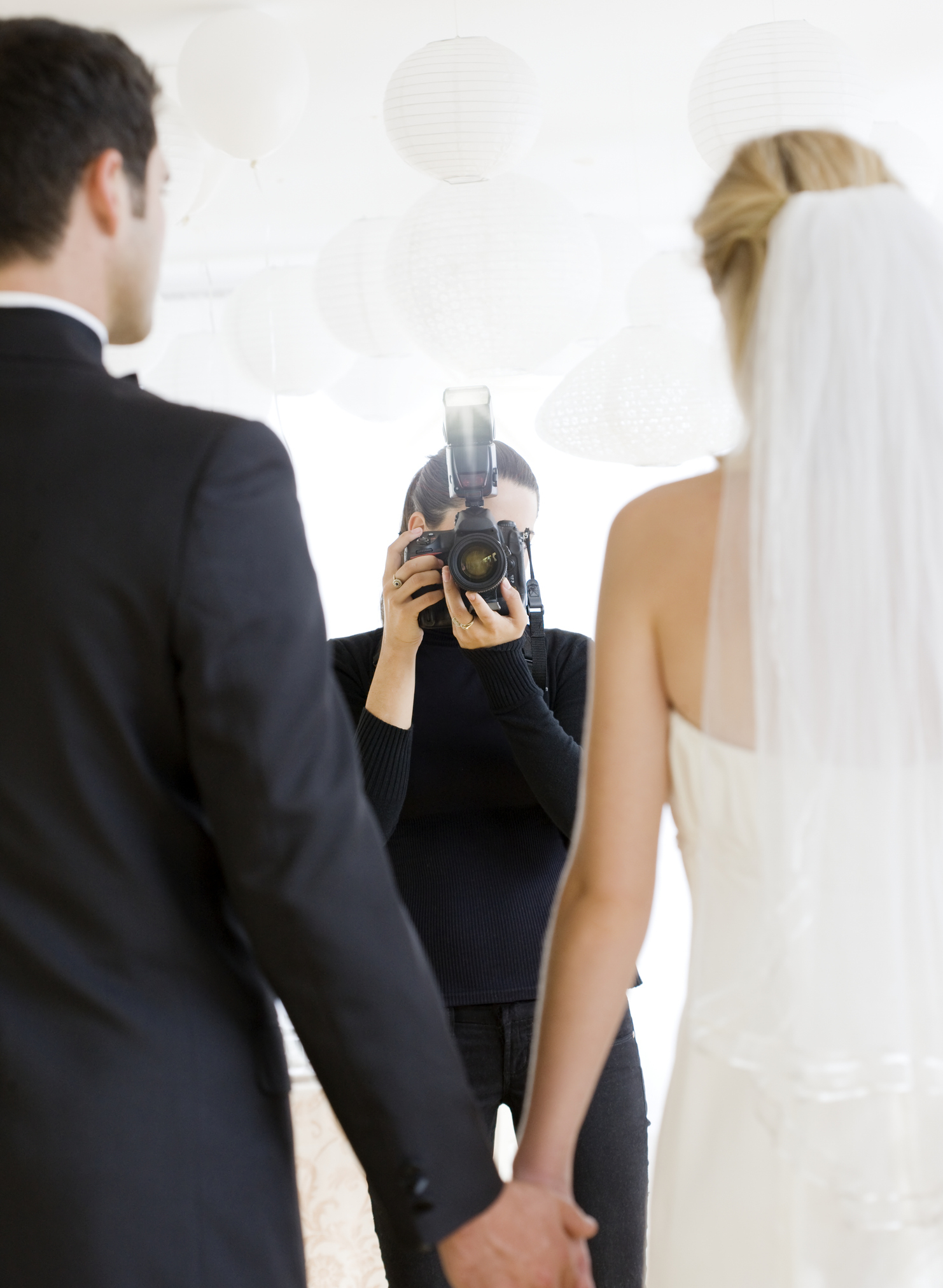 A bride and groom hold hands while facing a photographer taking their photo. The bride wears a wedding dress and veil; the groom is in a suit