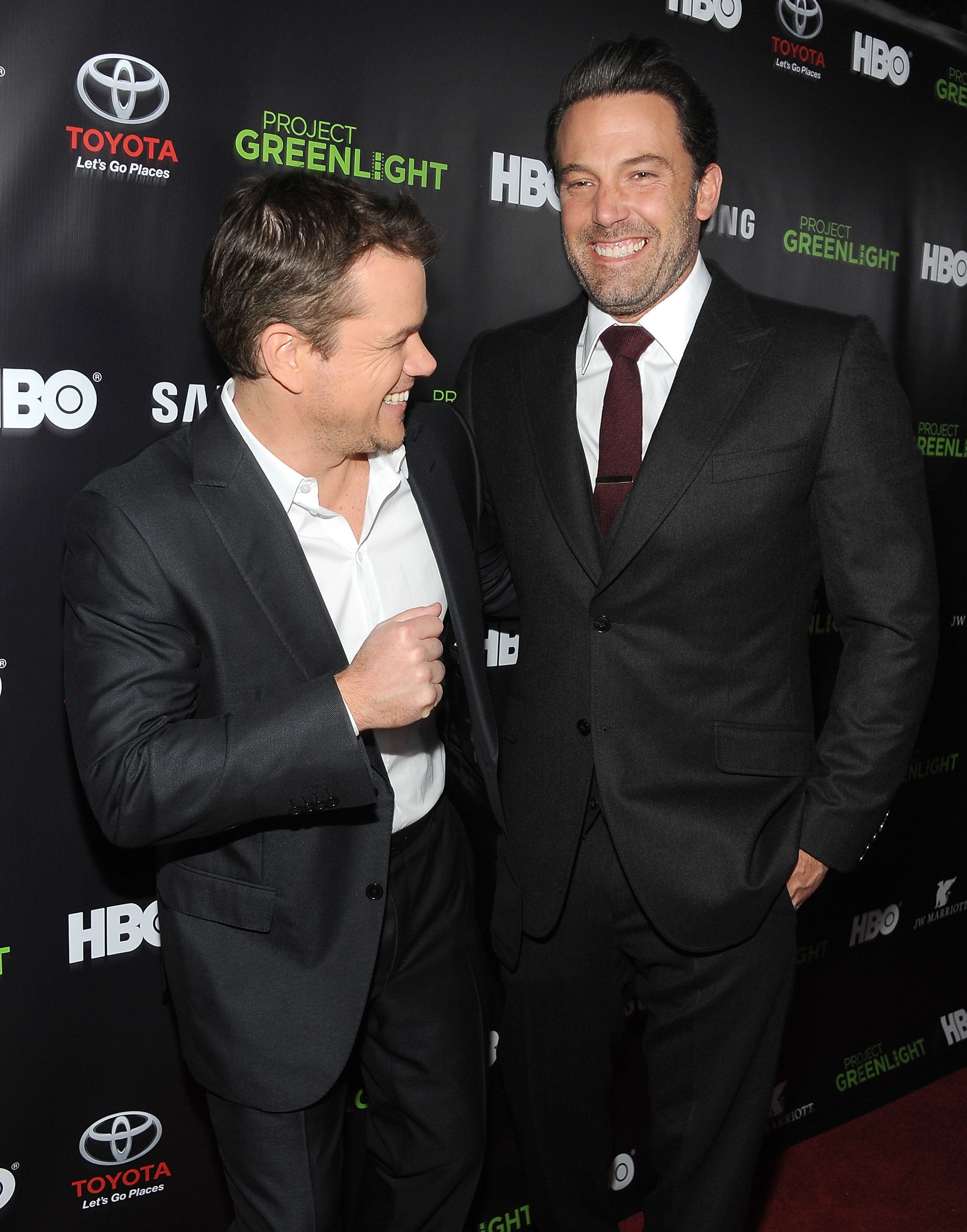Matt Damon and Ben Affleck on the red carpet at a Project Greenlight event, both wearing suits and smiling at each other