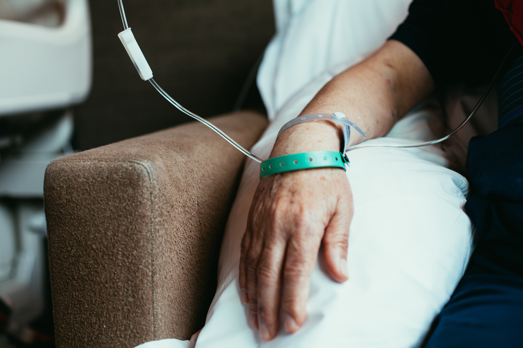 A close-up of an unidentified person's arm with an IV drip and hospital wristband, resting on a pillow on a hospital bed