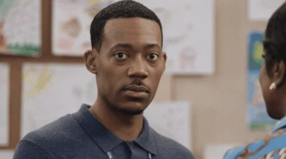 A person is looking directly at the camera, standing in front of a display of drawings on a wall. Other person in foreground's shoulder is partially visible