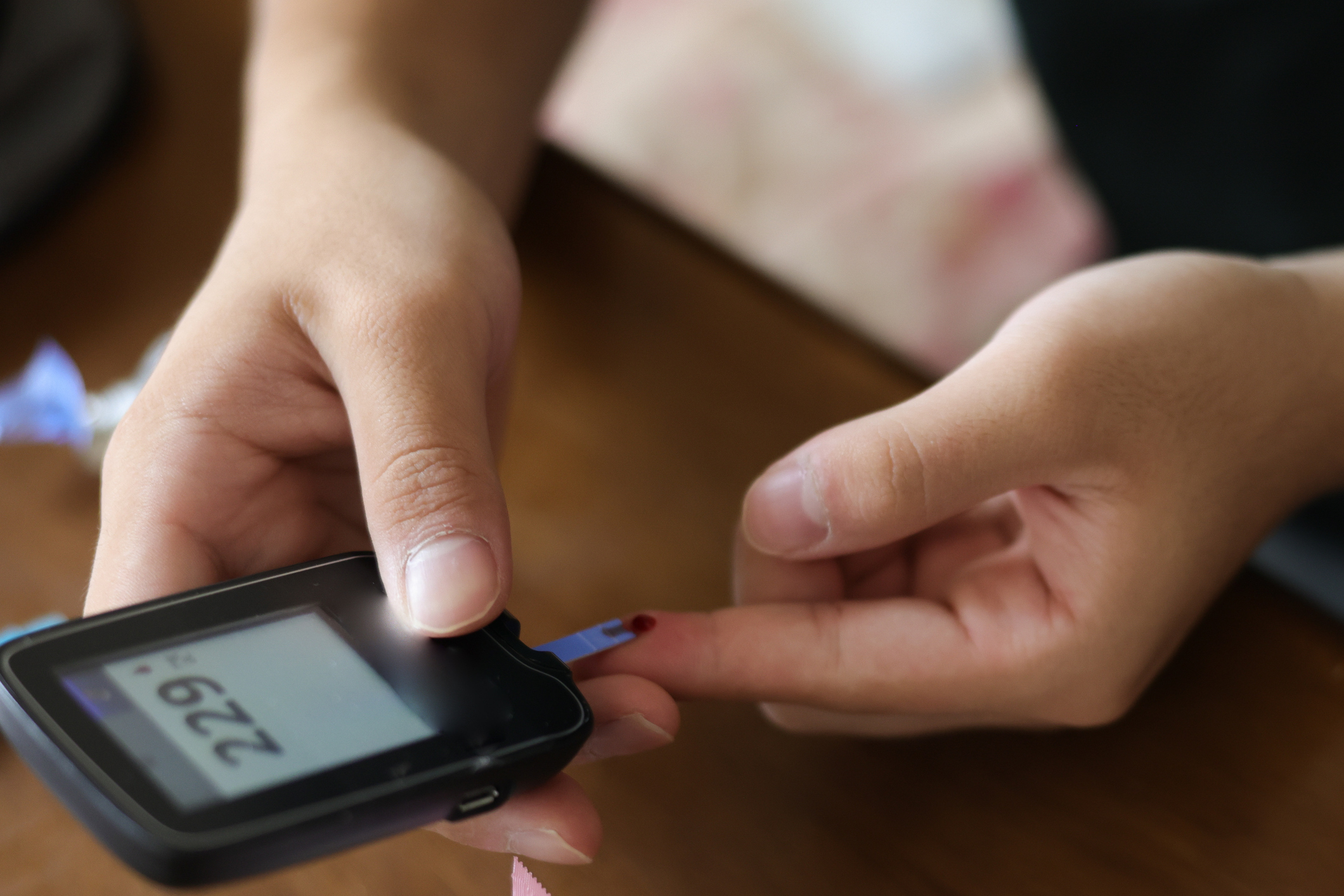 A person tests their blood sugar level using a glucose meter, which displays a reading of 229. A small blood droplet is on one finger