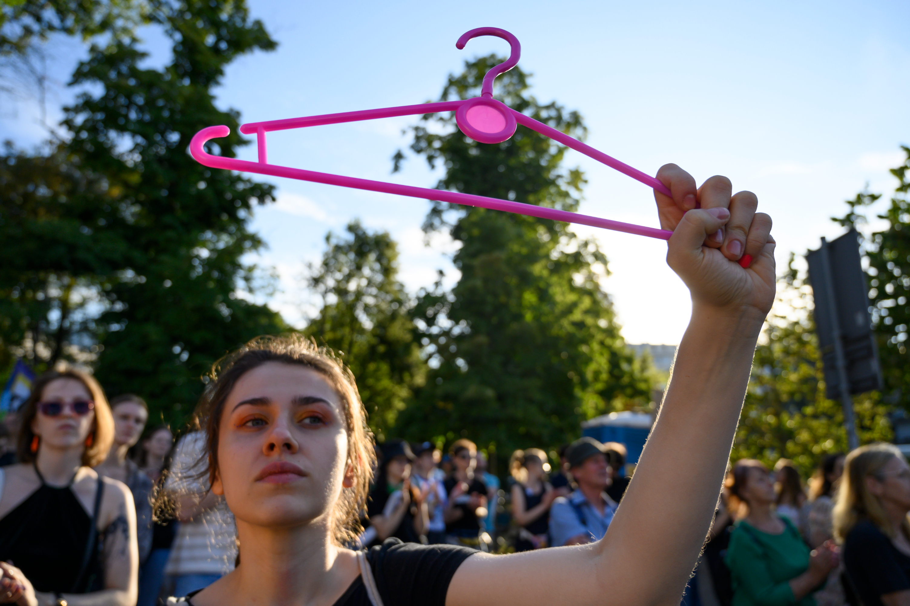 A woman in a crowd holds up a pink hanger at an outdoor gathering, indicating a protest. Other participants are visible in the background