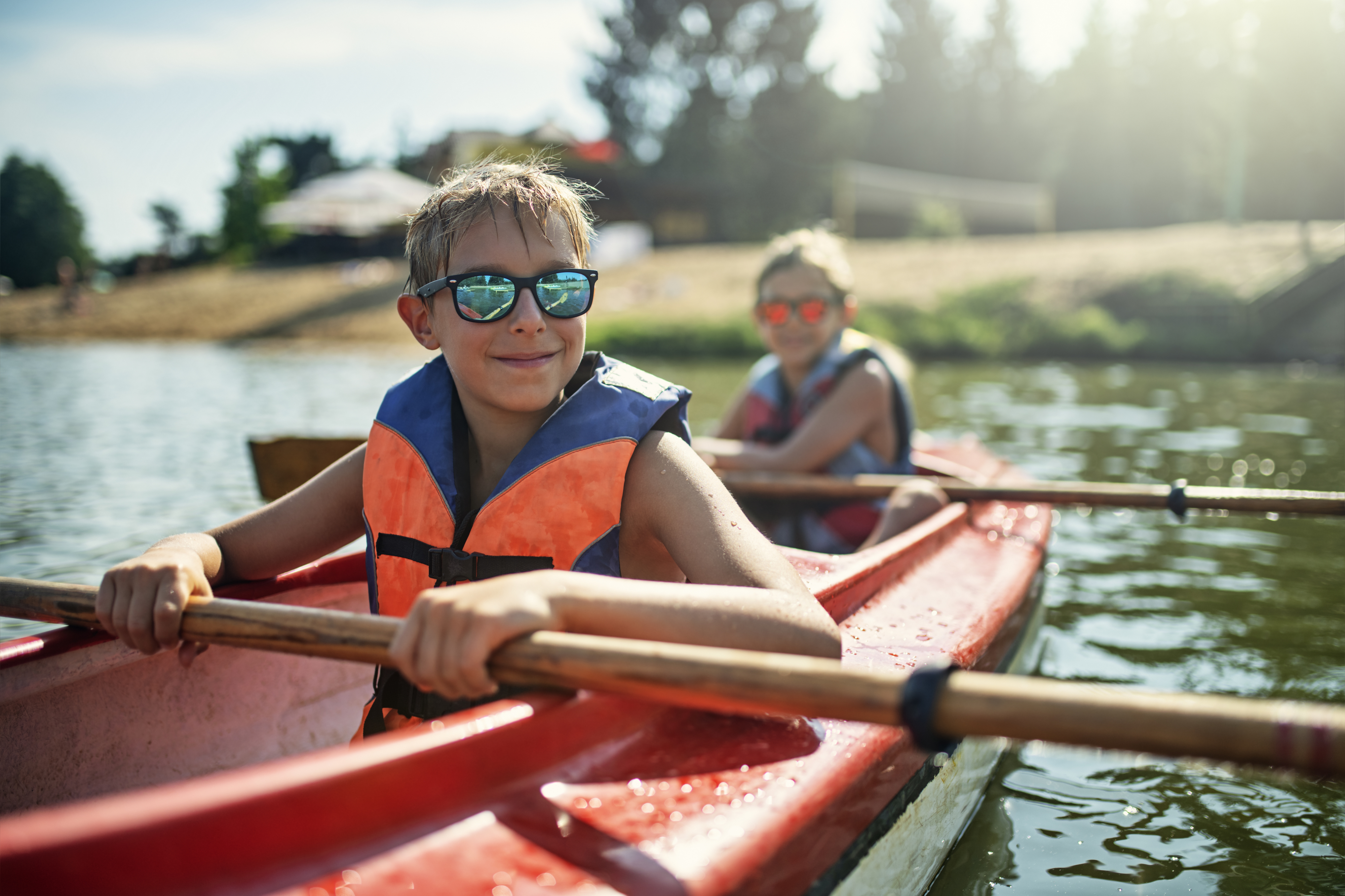 Two children are kayaking on a calm lake, both wearing life jackets and sunglasses. They are paddling and smiling toward the camera, enjoying a sunny day