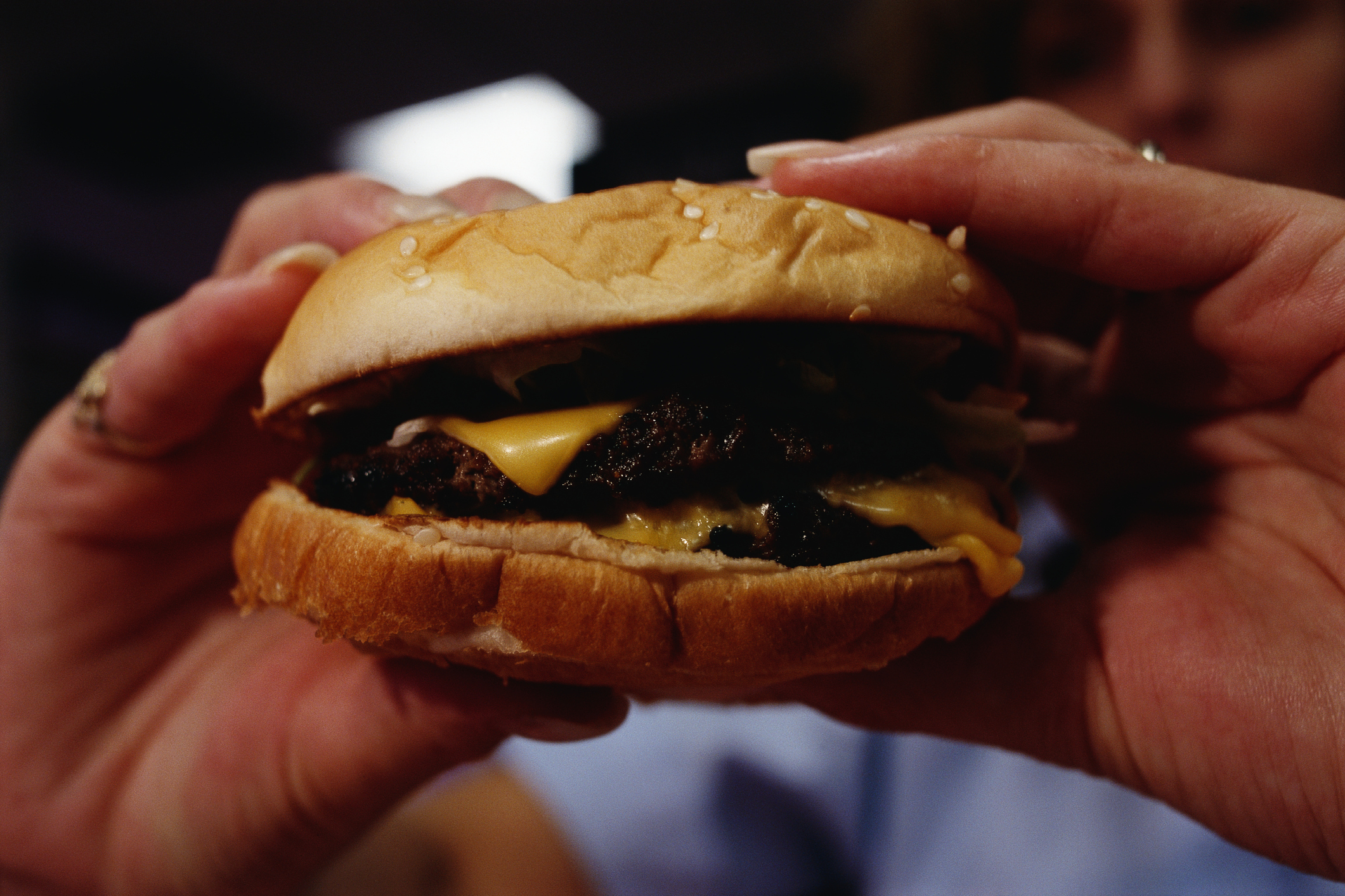 A person holding a cheeseburger with their hands, showcasing the burger's sesame seed bun, melted cheese, and beef patty. The person's face is mostly out of view