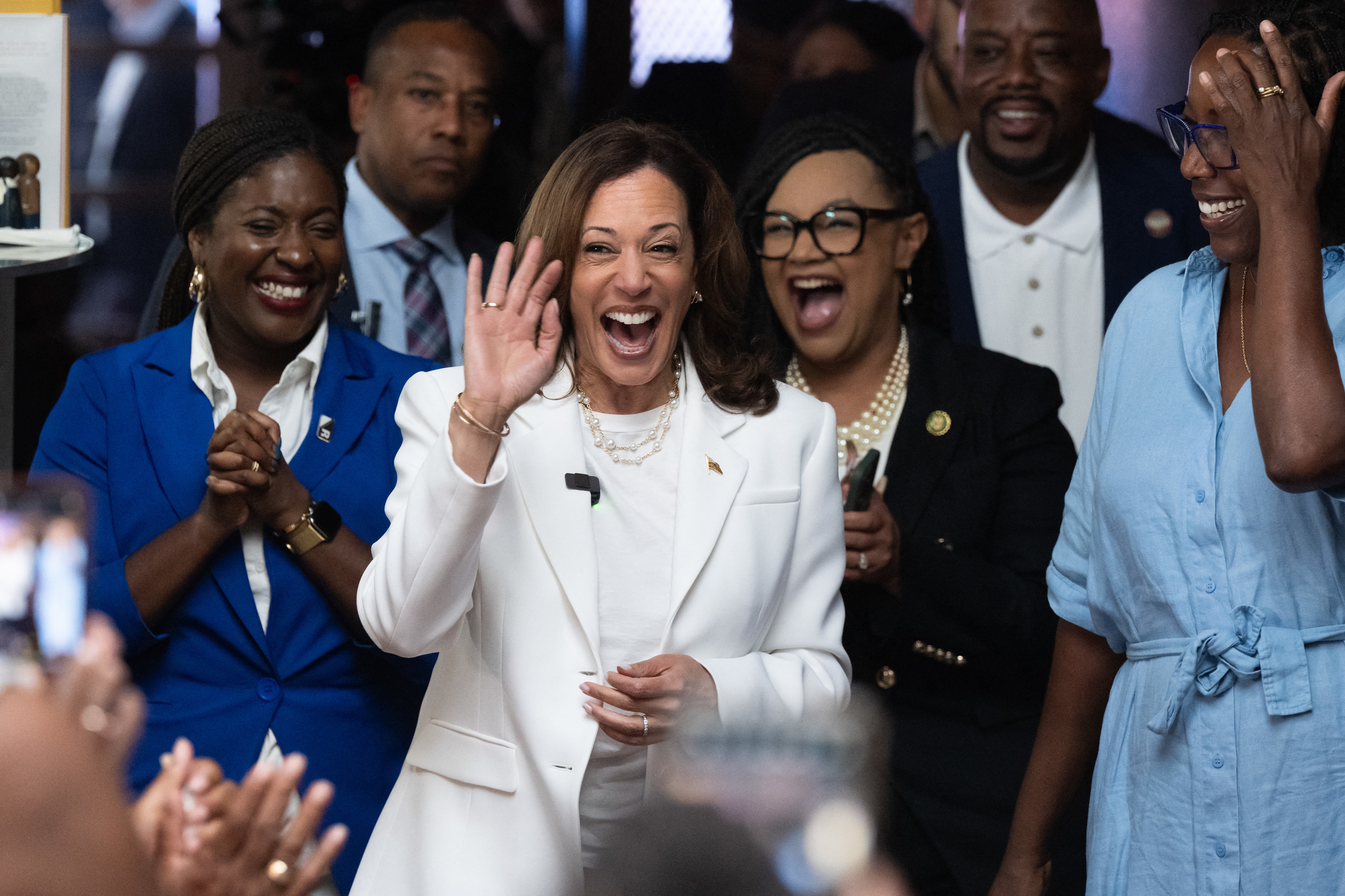 Kamala Harris, in a white suit, waves and smiles while surrounded by a group of people, some clapping and cheering, in an indoor setting