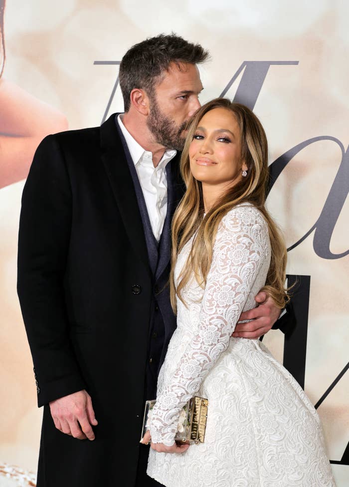 Ben Affleck in a dark coat kisses Jennifer Lopez, who is in a white lace dress, on her head while they pose together at a public event