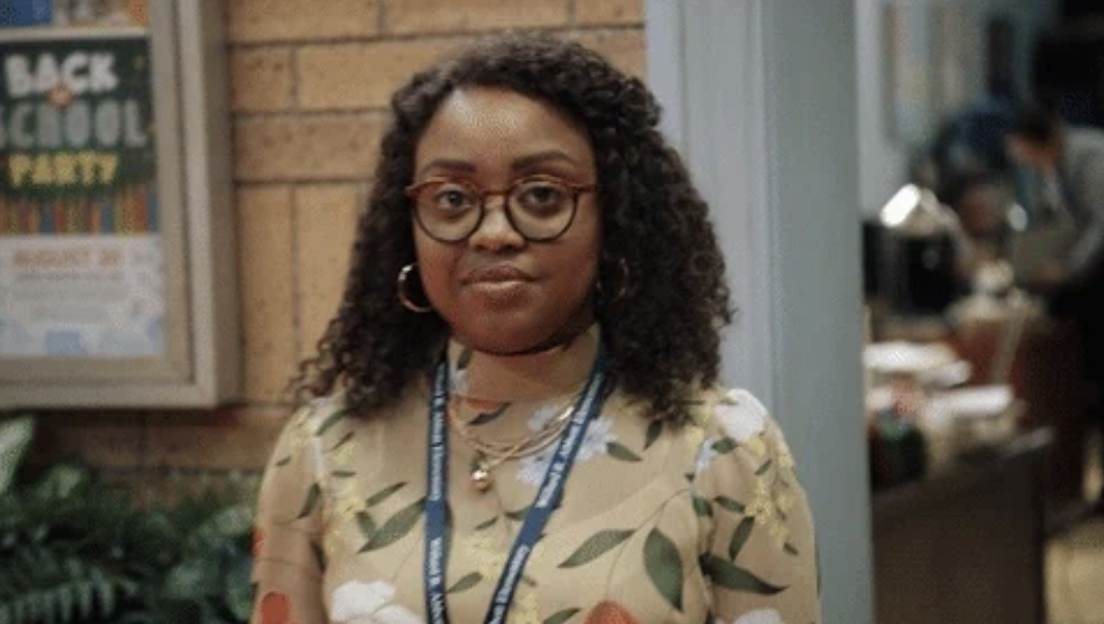 Quinta Brunson stands in an indoor setting, wearing a floral-patterned blouse and glasses, with a lanyard around her neck. There is a "Back to School Party" poster behind her