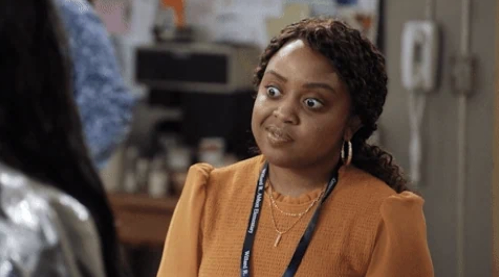 Quinta Brunson, standing indoors, wearing a long-sleeved blouse and a lanyard, with a humorous and wide-eyed expression on her face