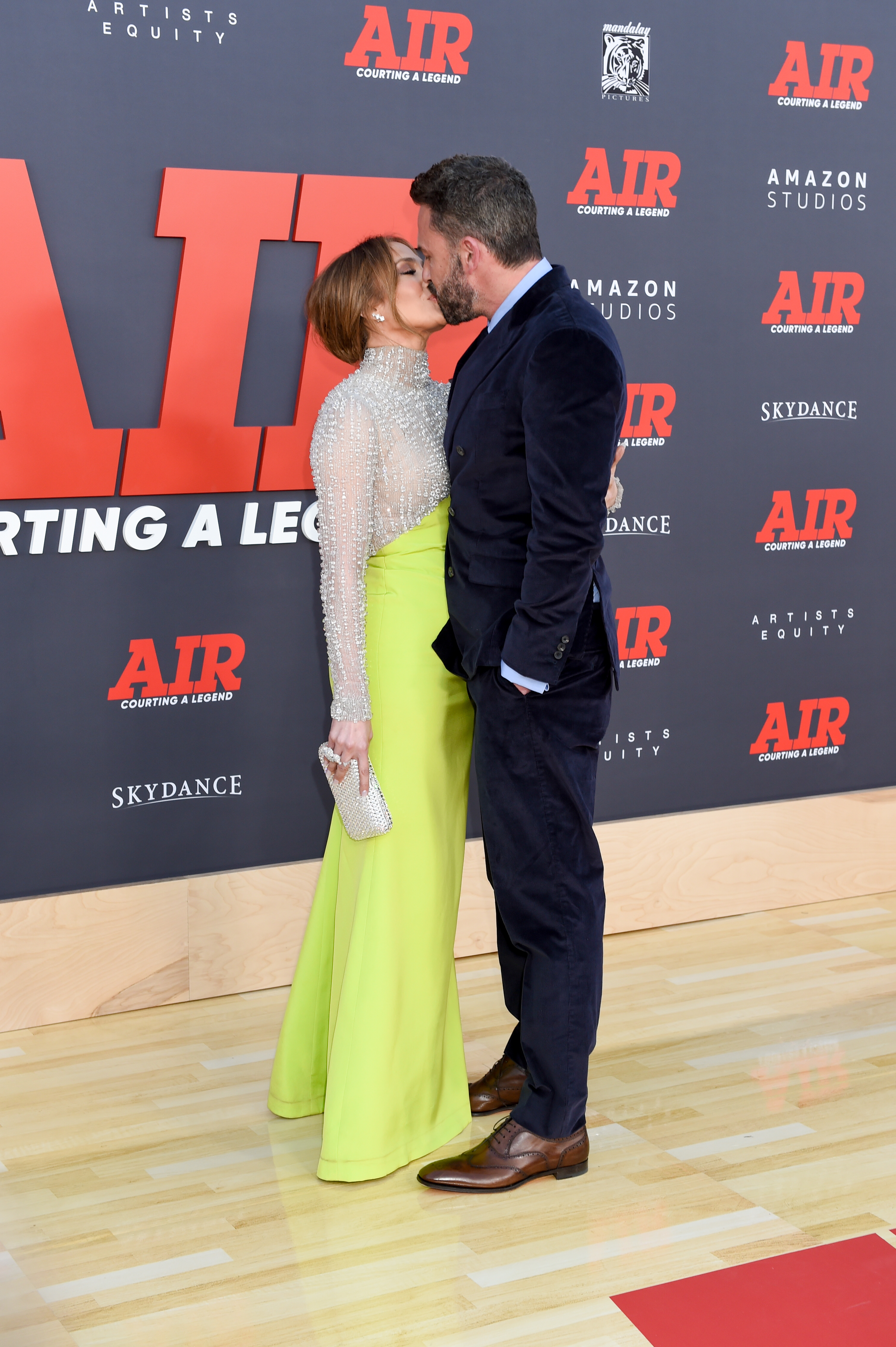 Jennifer Lopez in a high-neck, long-sleeve bodice with a neon green skirt kisses Ben Affleck in a navy velvet suit on the red carpet at the 'Air' premiere