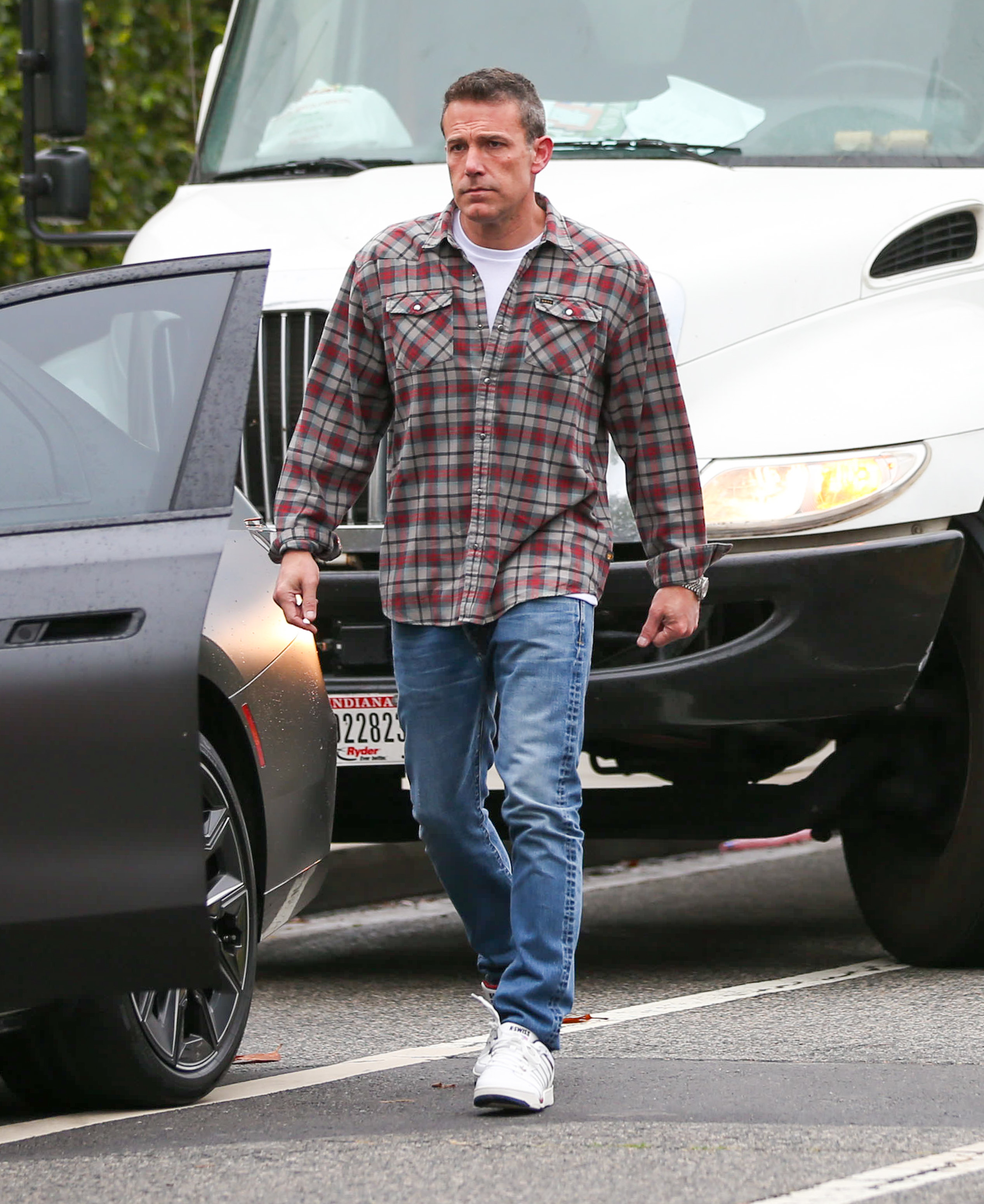 Ben Affleck walks across a street wearing a checked flannel shirt, jeans, and white sneakers. A car door is open next to him