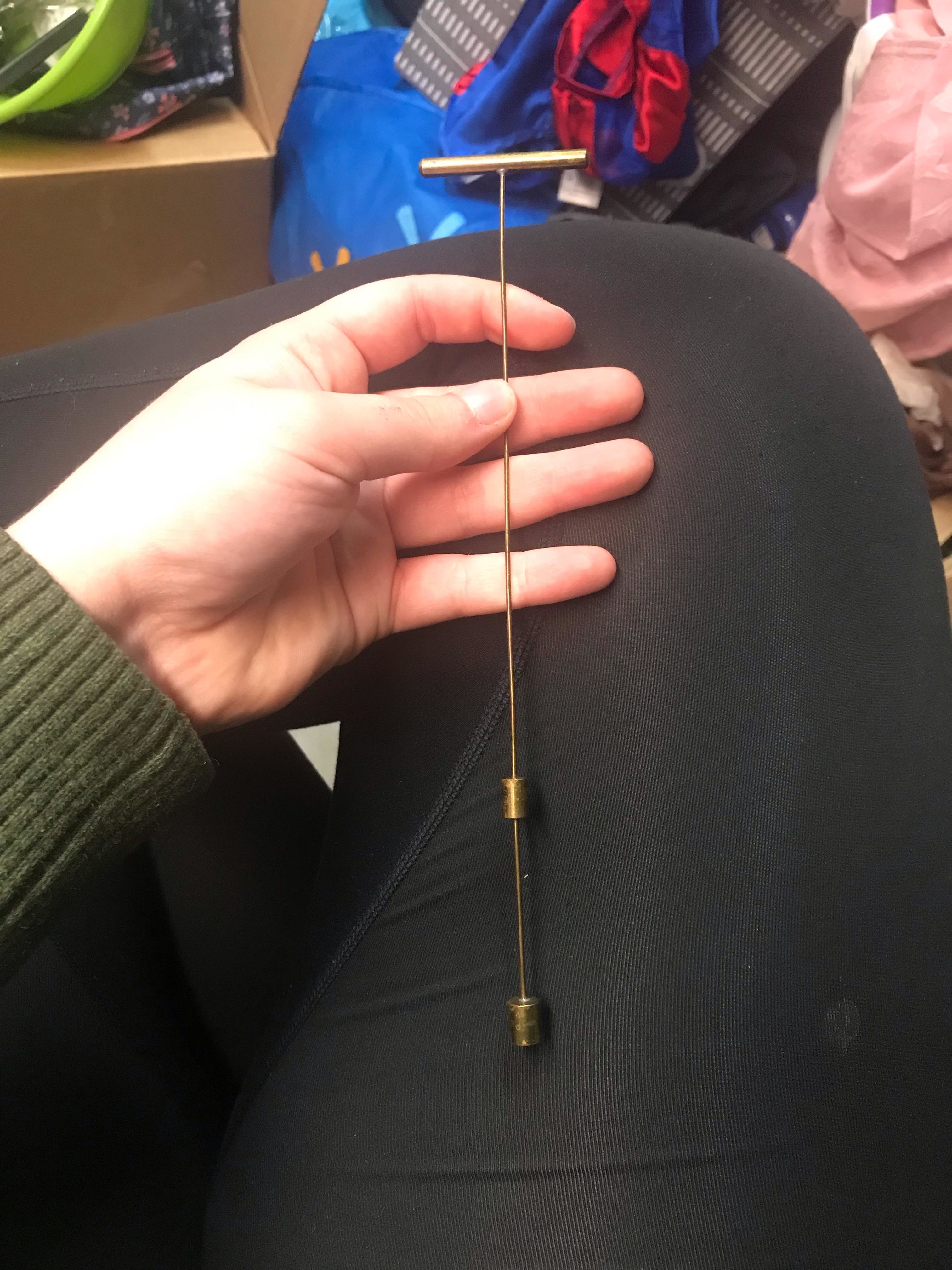 A hand holding a long, thin metal tool resembling a T-shaped rod with two adjustable components. The background contains miscellaneous items