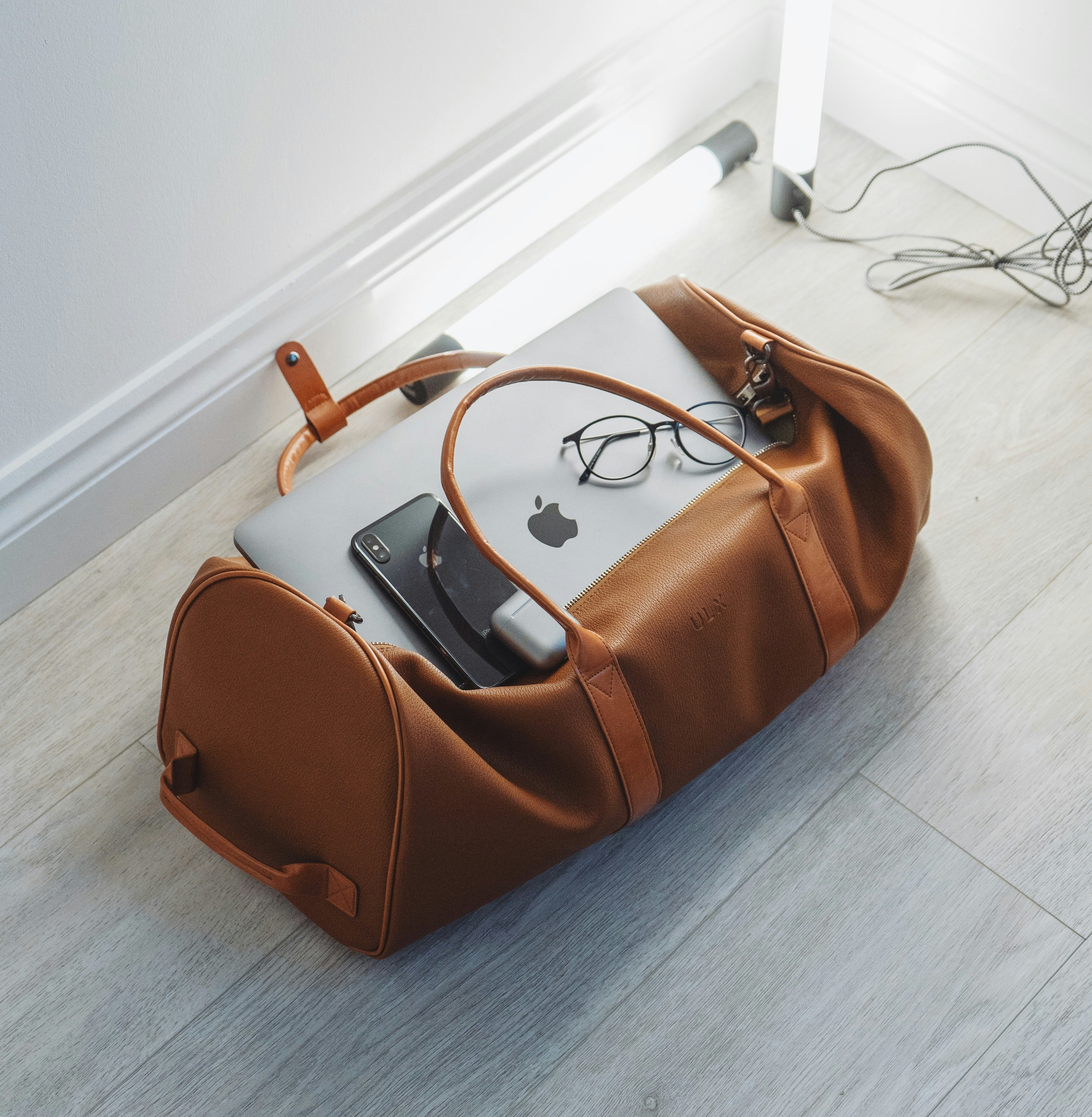 A brown leather duffel bag on the floor holds a MacBook, a pair of eyeglasses, and various electronic accessories