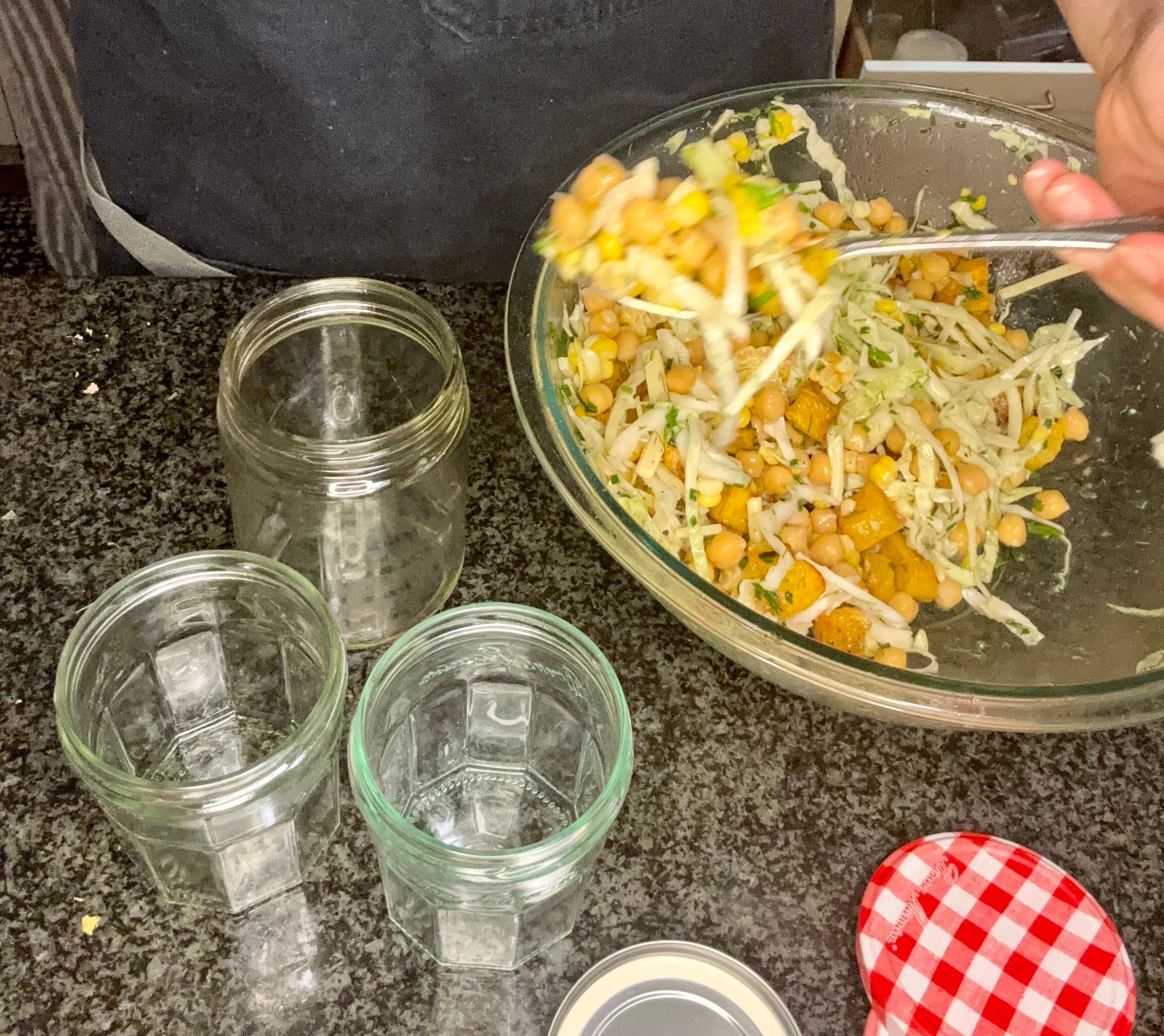 Person wearing a dark apron and green shirt preparing a salad with chickpeas and vegetables, holding a spoon above glass jars on a countertop
