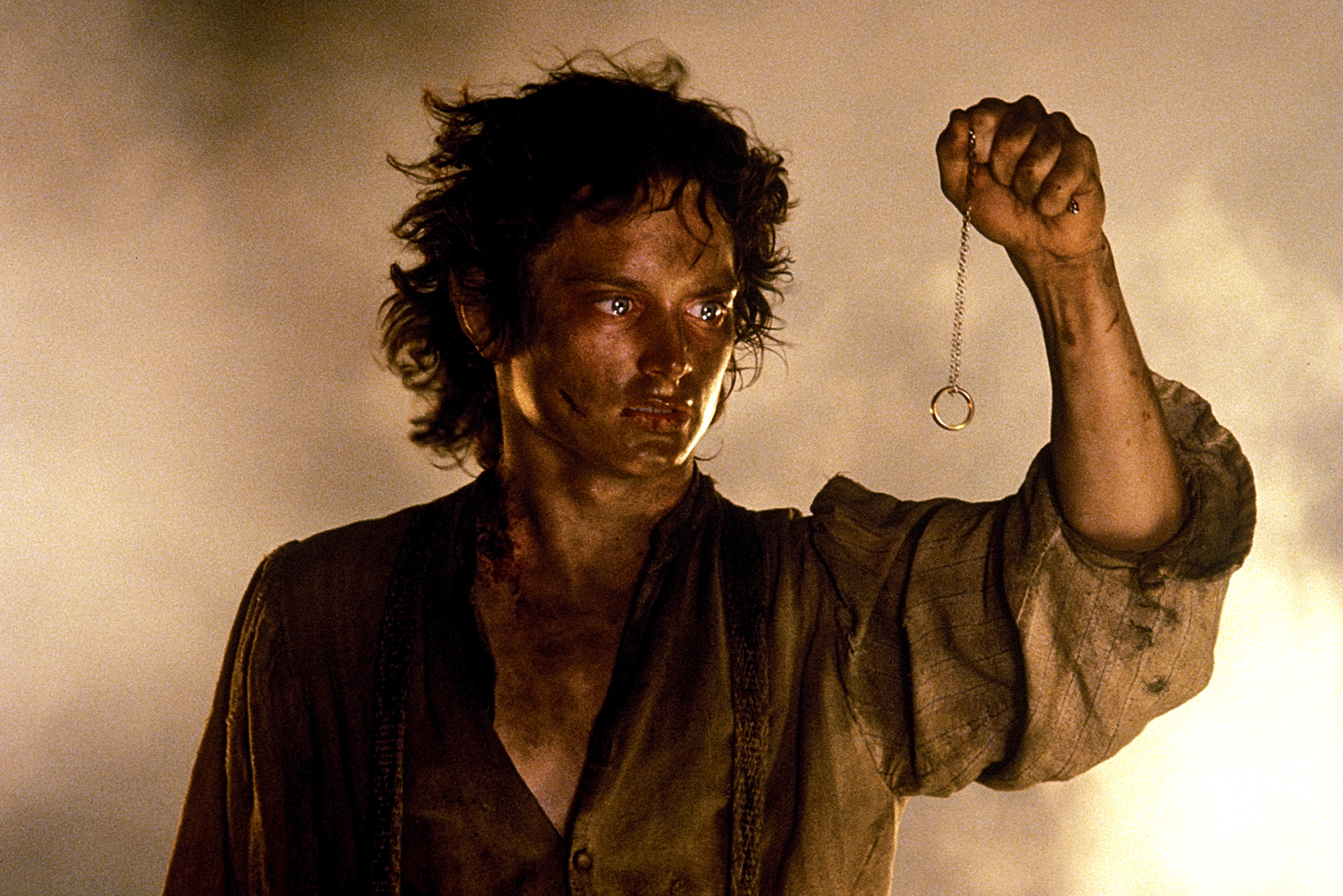 Elijah Wood as Frodo Baggins from "The Lord of the Rings" holds the One Ring on a chain, with a determined and worn-out expression