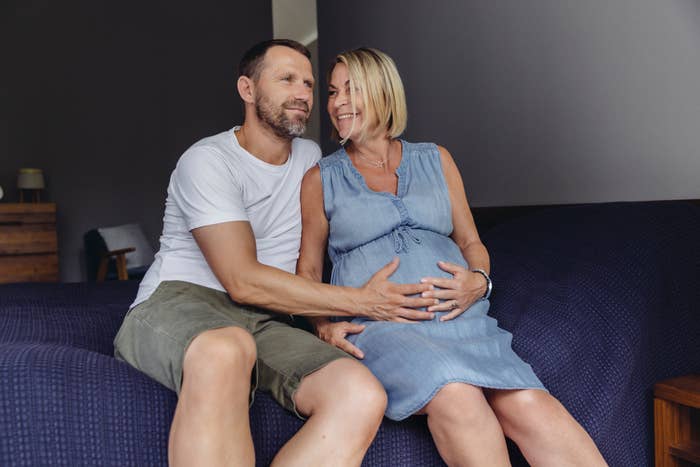 An expectant couple sitting on a bed, the man has his hand on the woman's pregnant belly