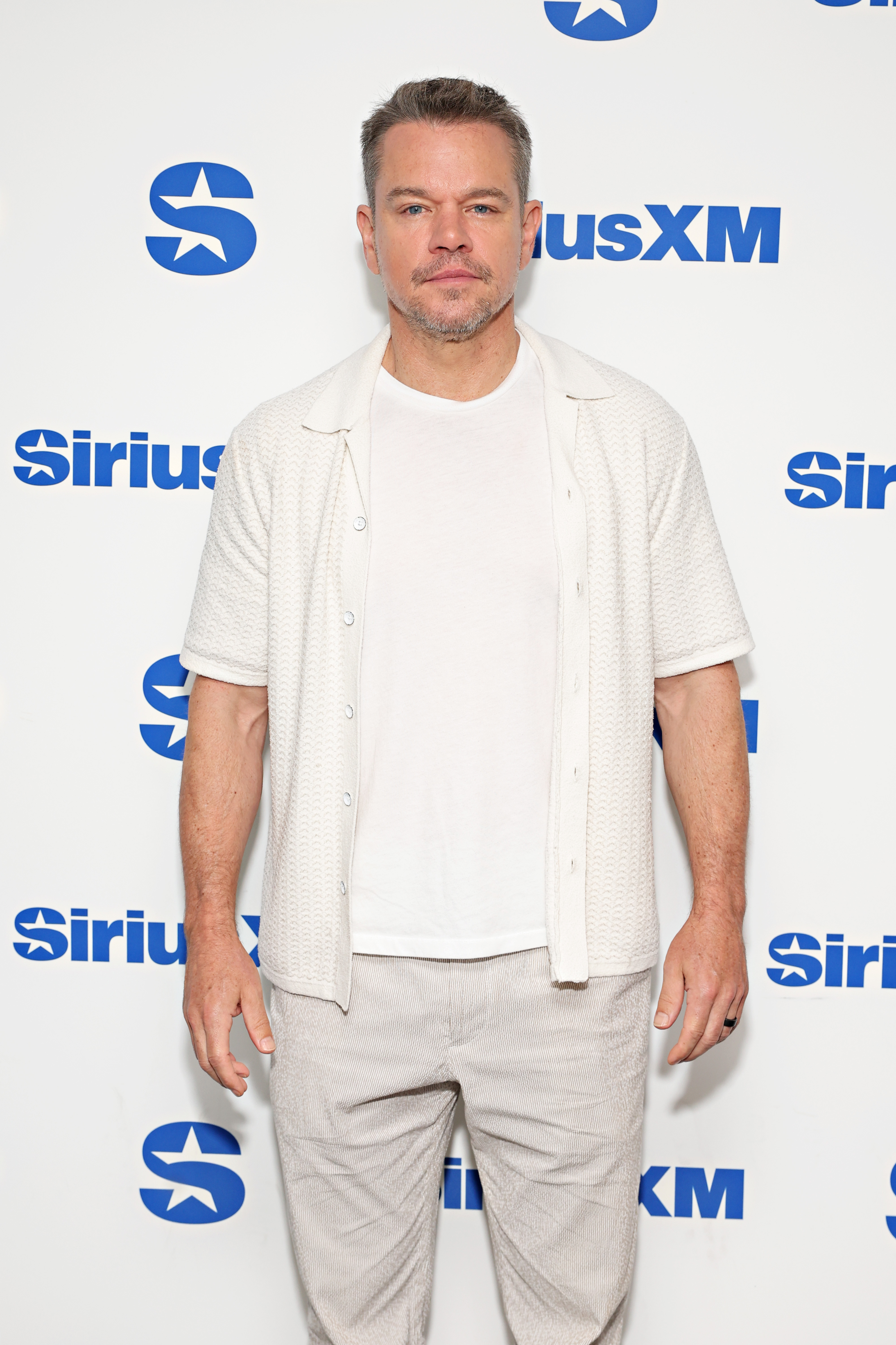 Matt Damon at a SiriusXM event, wearing a casual white shirt over a white T-shirt and light-colored pants, standing in front of a SiriusXM branded backdrop