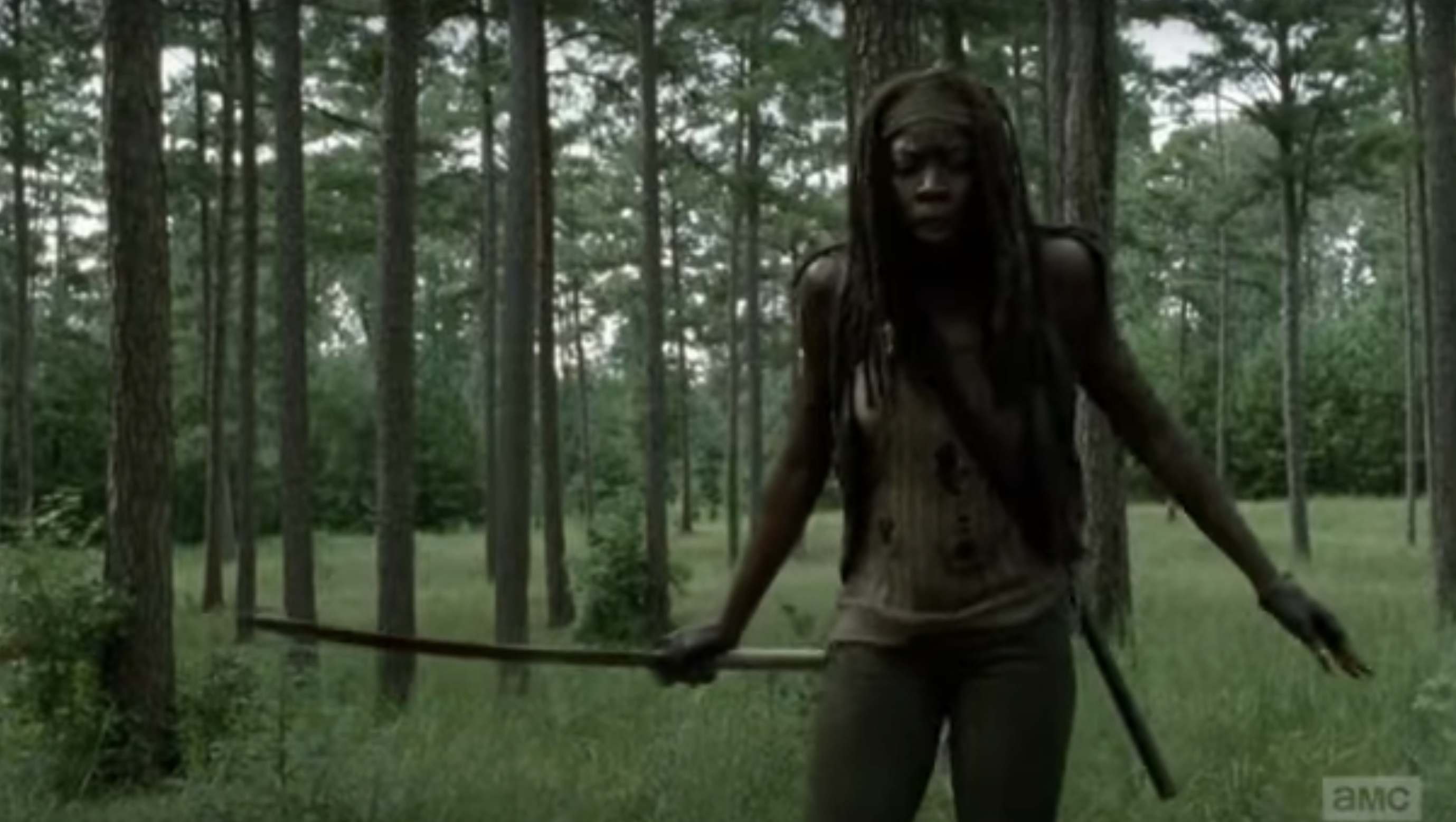 Danai Gurira as Michonne from The Walking Dead stands in a forest holding her katana, looking intensely ahead