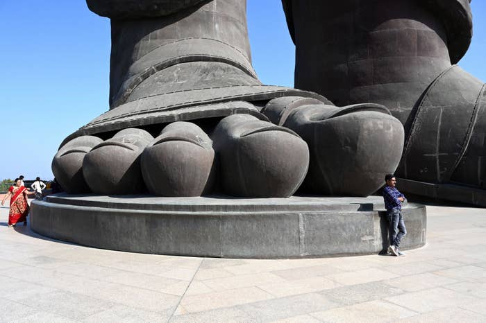 People pose near the enormous feet of a giant statue. A woman in a sari on the left, and a man in casual clothing on the right lean against the statue