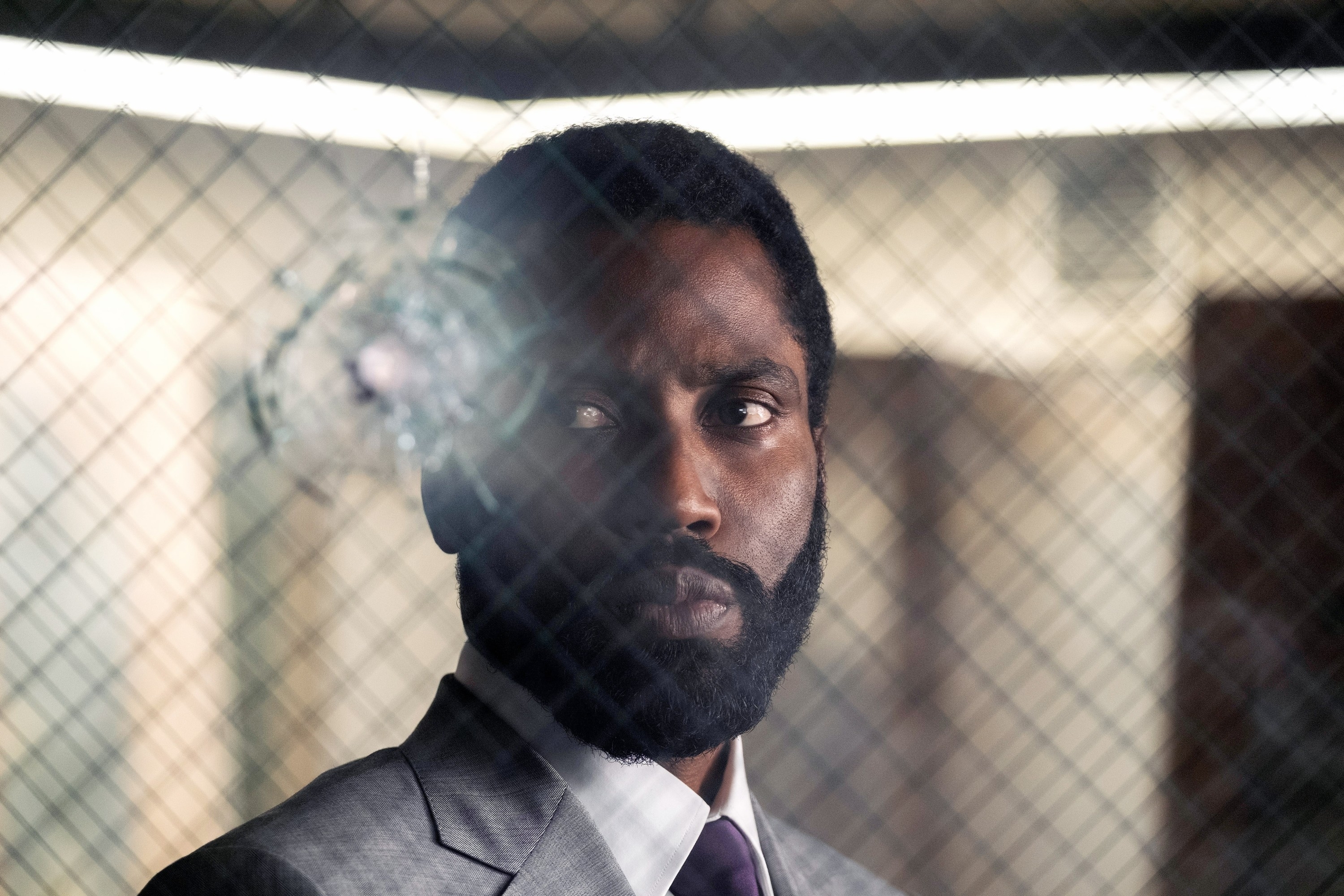 John David Washington in a suit stares intensely through a glass window with a bullet hole crack, hinting at a suspenseful scene