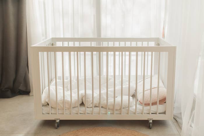 An empty white baby crib with bedding and a small pillow, placed near a window with sheer white curtains
